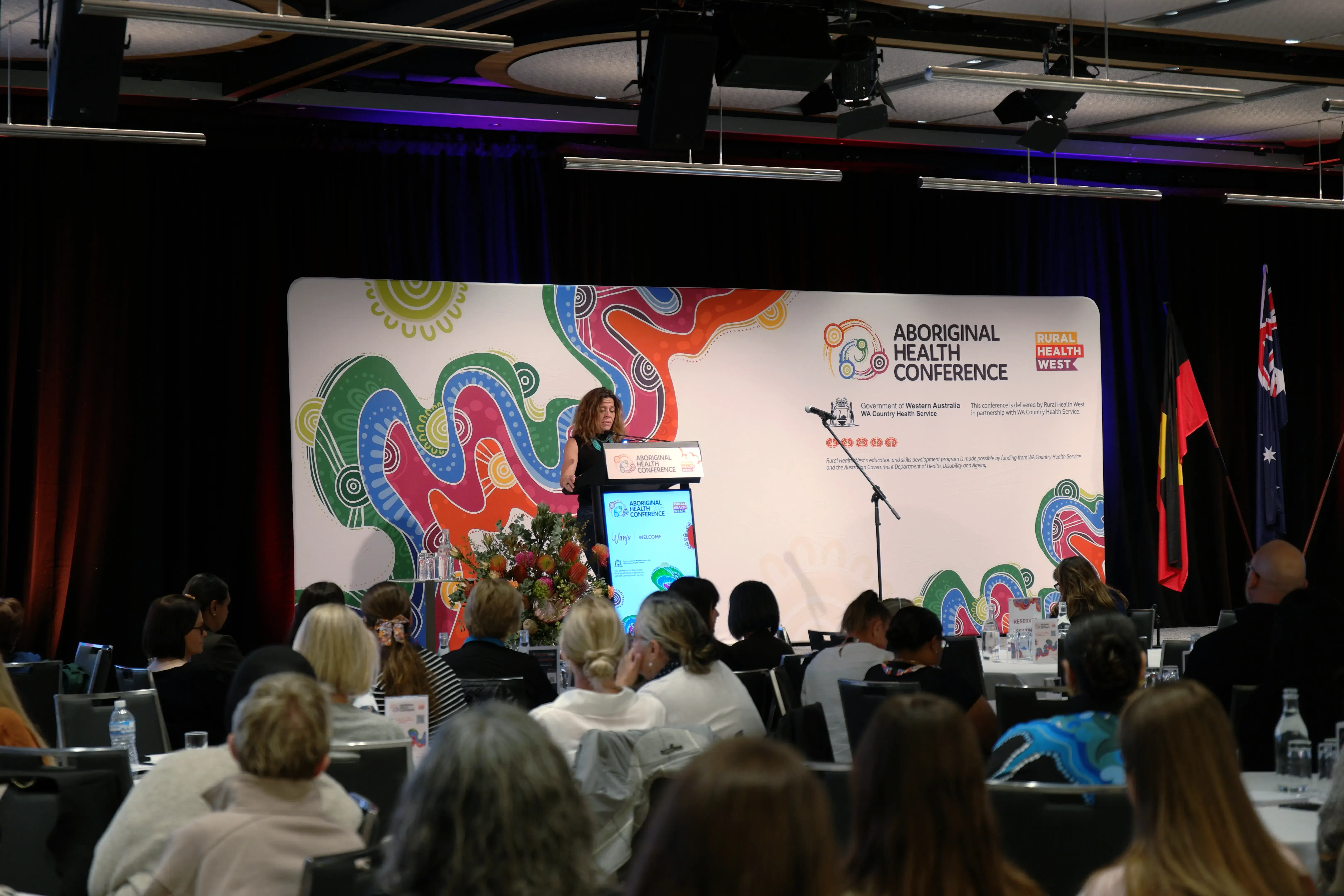 A speaker presenting on stage while delegates listen from their tables at the Aboriginal Health Conference 2026 in Perth.