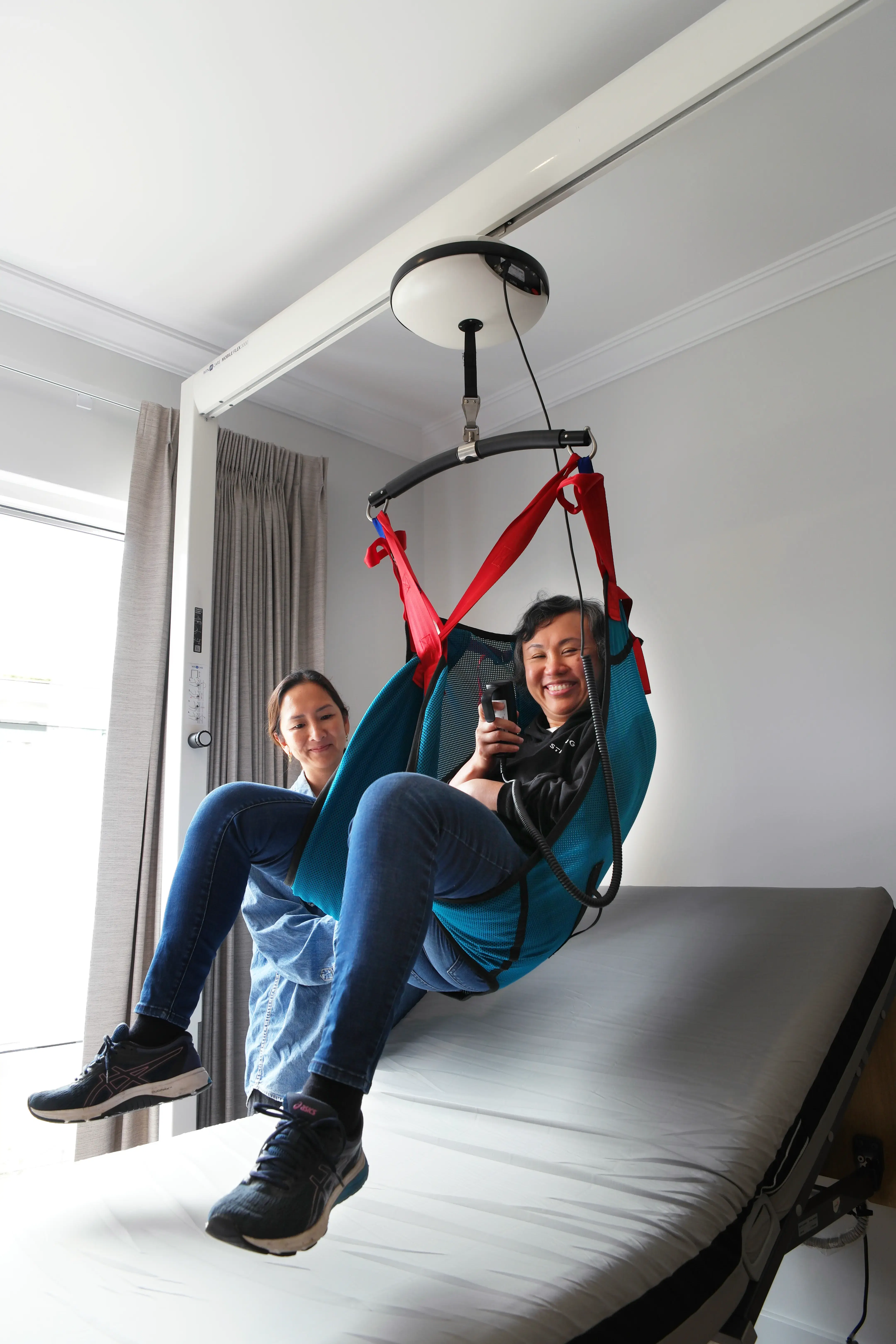 Person suspended in a red patient lifting sling during a ceiling track hoist demonstration above an adjustable care bed.