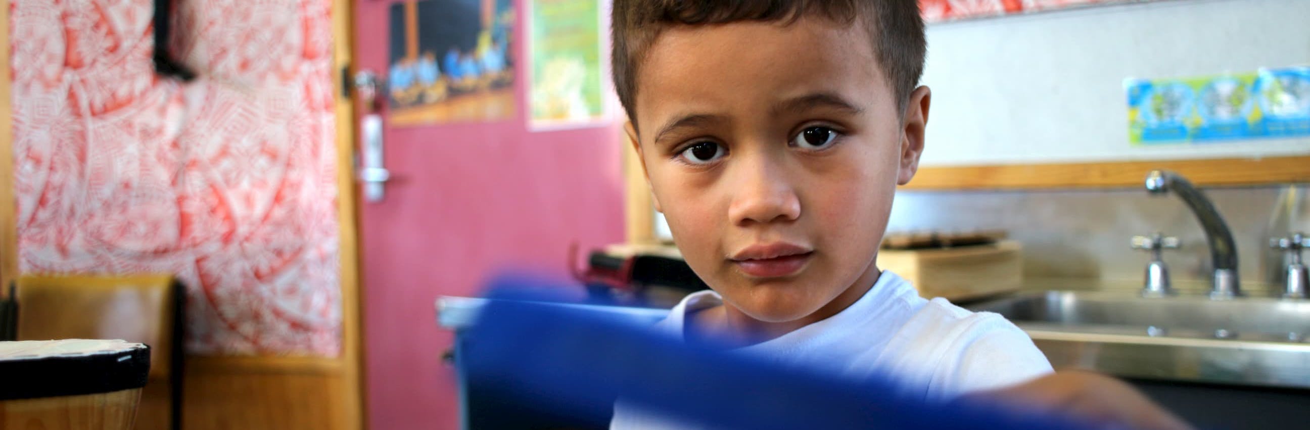 A young Māori boy looks at the camera while standing in a classroom