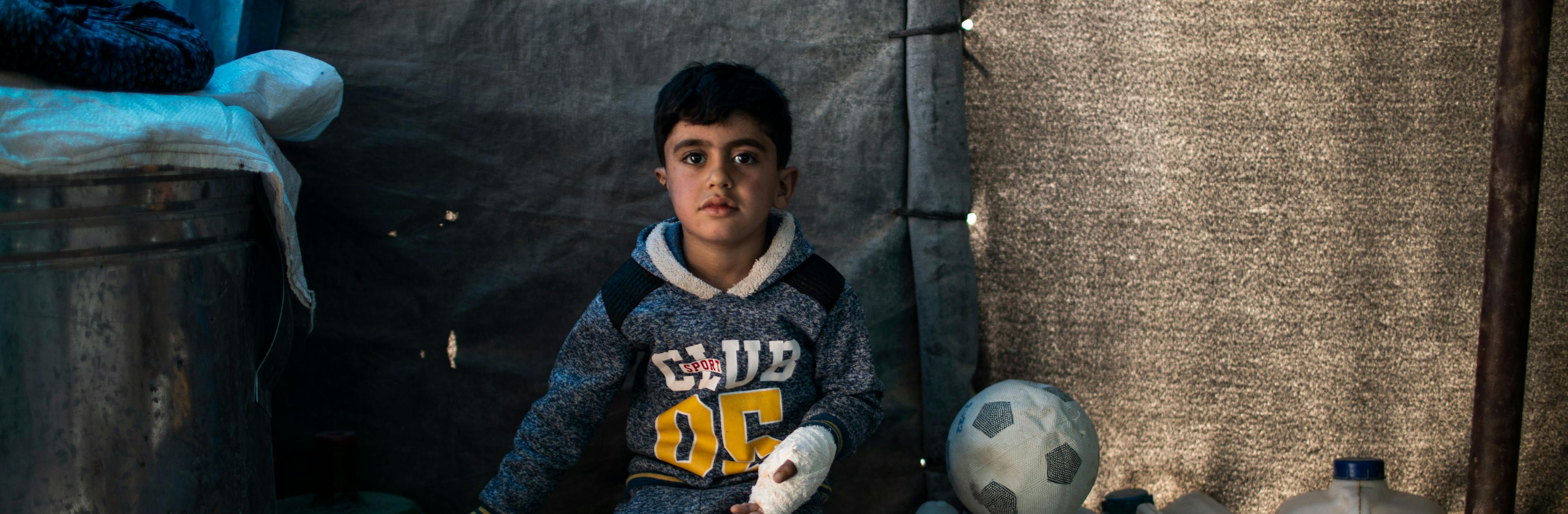 On 28 February 2022 in Iraq, 5-year-old Hussein sits on a water container at his temporary home in Habaniya, on the outskirts of Fallujah. In November 2021, Hussein was playing soccer with his brother when a piece of previously unexploded ordinance (UXO) detonated under him, ripping open his stomach and leaving part of his right ear missing.