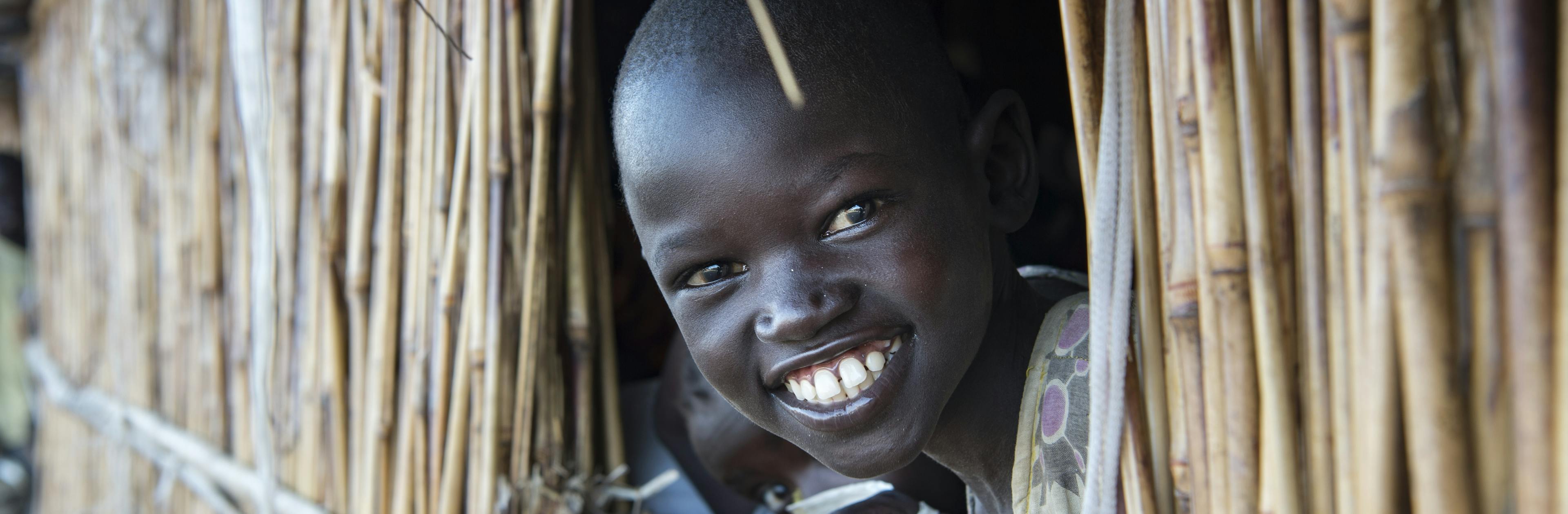 About Us- A young girl smiles as she looks out a classroom window at Lich Primary School, located at the Bentiu Protection of Civilians (POC) site, at the UNMISS (United Nations Mission in South Sudan) base