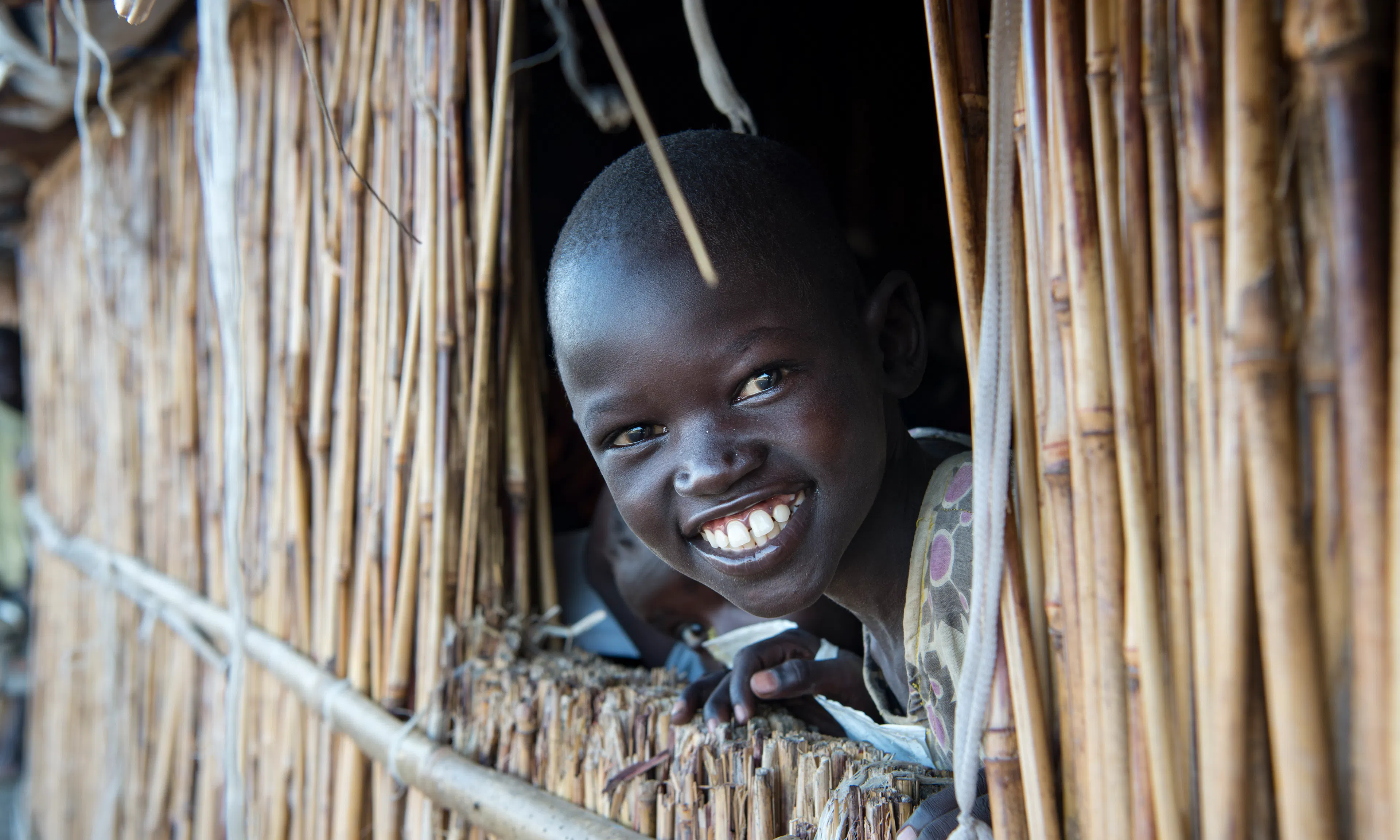 About Us- A young girl smiles as she looks out a classroom window at Lich Primary School, located at the Bentiu Protection of Civilians (POC) site, at the UNMISS (United Nations Mission in South Sudan) base