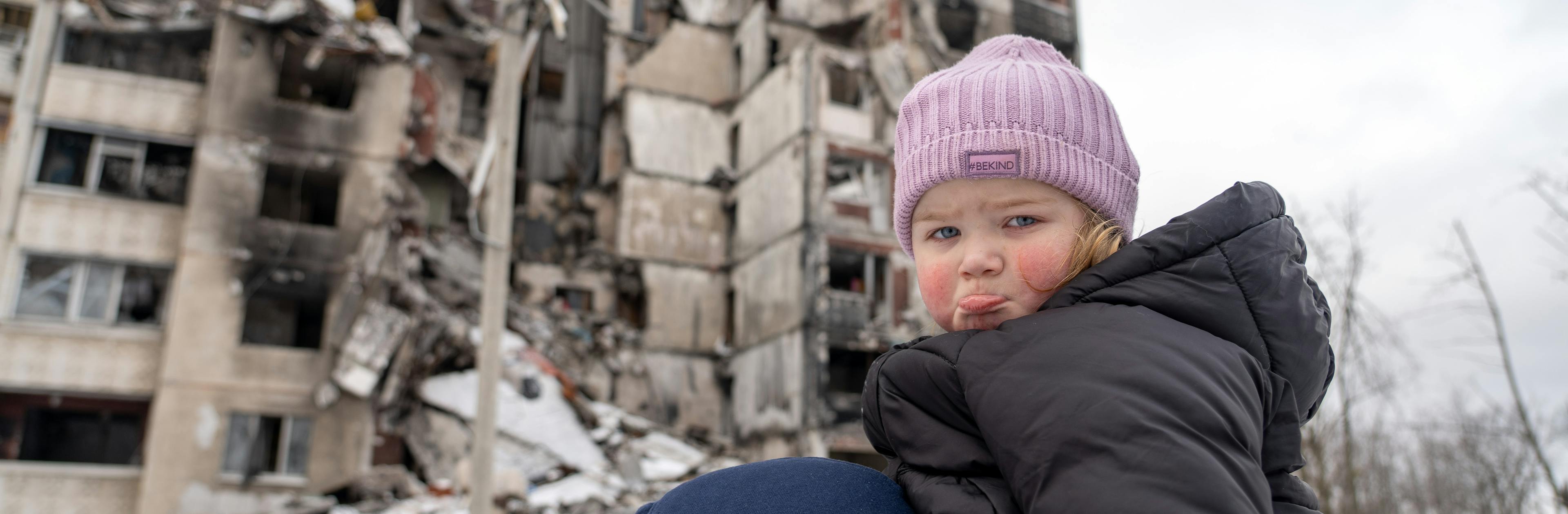 Little girl looks sad at the camera in front of a destroyed building in Ukraine
