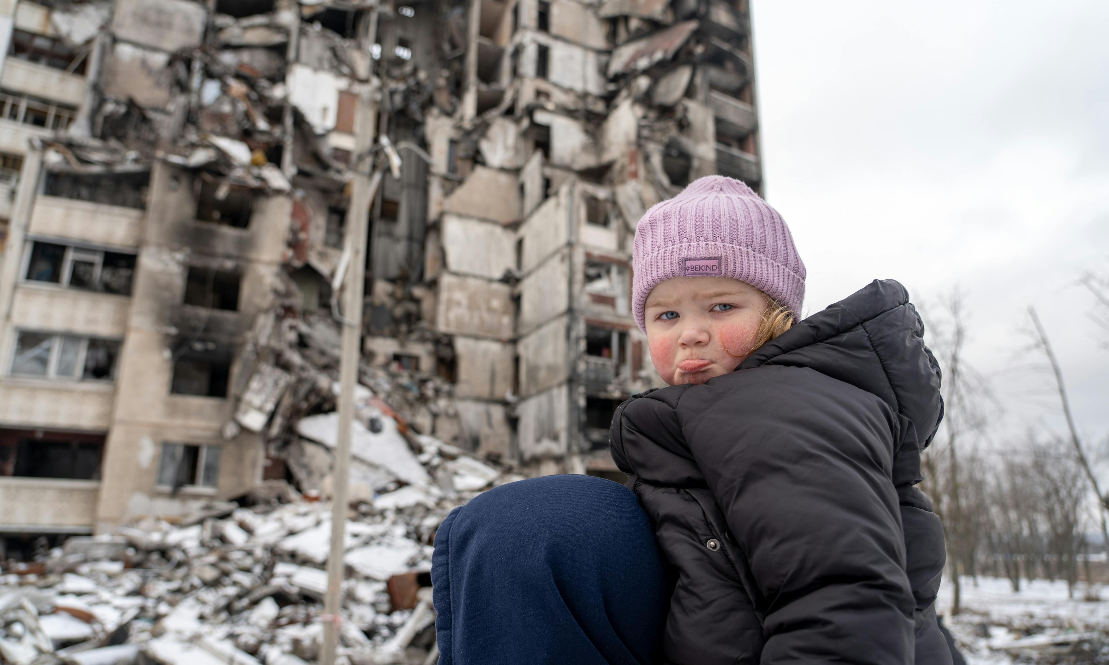 Little girl looks sad at the camera in front of a destroyed building in Ukraine