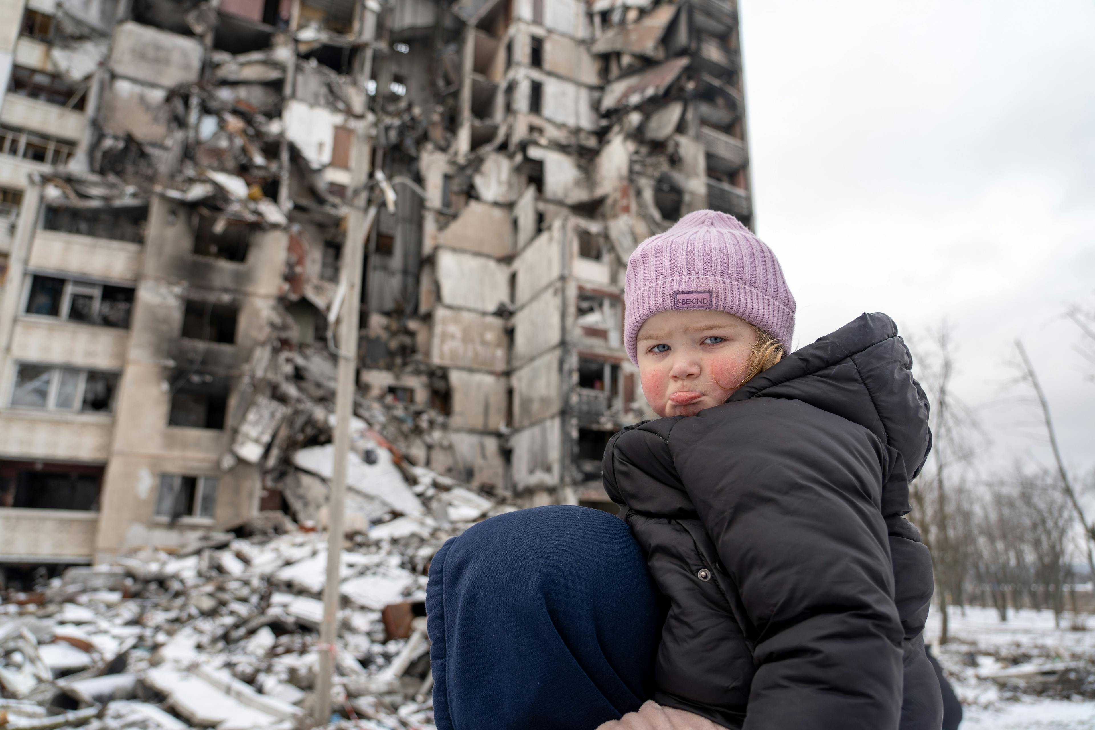 Little girl looks sad at the camera in front of a destroyed building in Ukraine