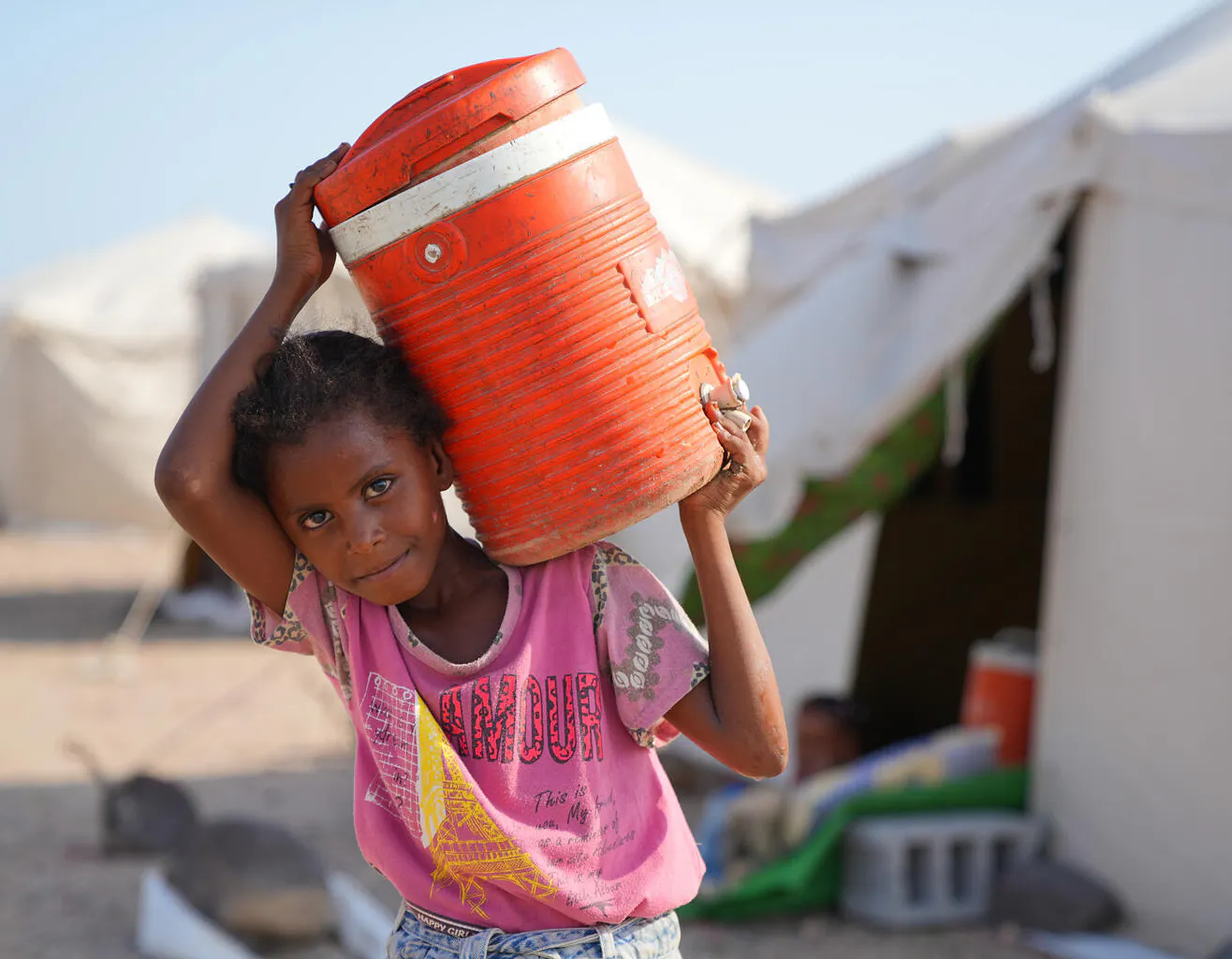 A young displaced girl carries a large orange water container on her shoulder at the Al-Firdous IDP camp in Aden, Yemen.