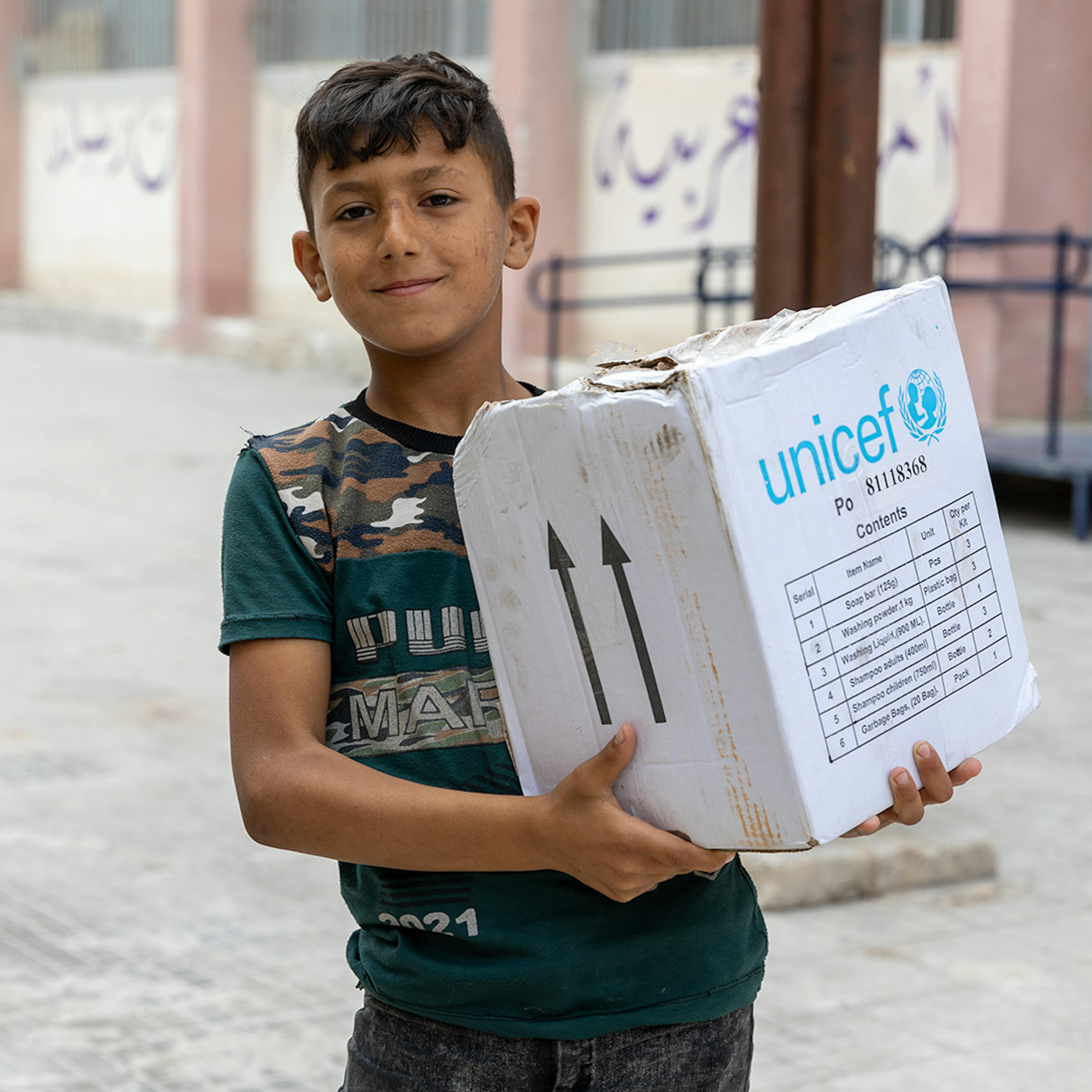 Abed, 10, carries his family hygiene box, distributed by UNICEF, at Bader Shaker Al Sayeb school-turned shelter in Aleppo city, Syria, on 14 June 2023.