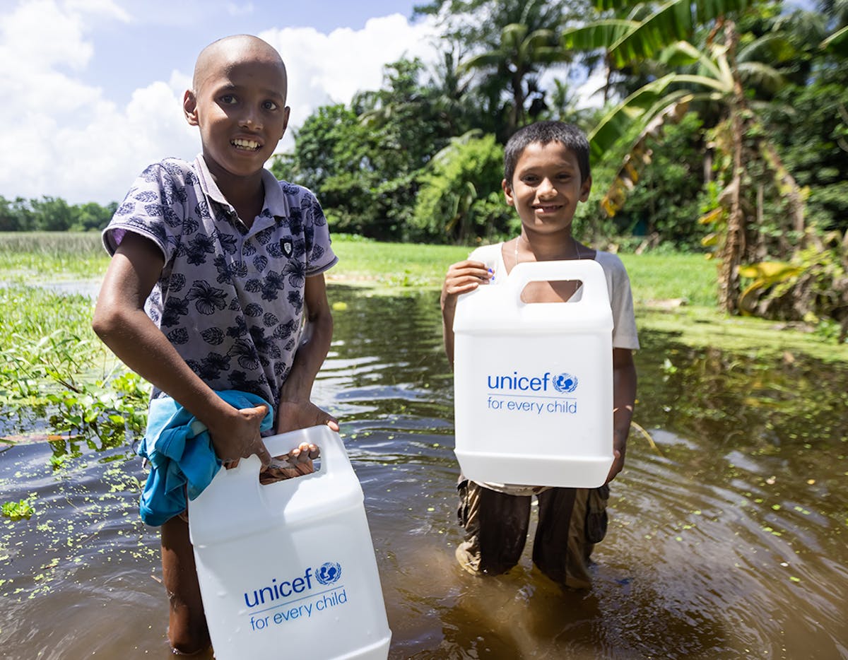 On 9 September 2024 in Badhitola Bazar, Kanchanpur, and Kadir Hanif Upazila, Bangladesh, Romiz, Kabir, and Shayal, who lost their home to the floods, carry a gallon of purified water distributed by UNICEF Bangladesh.