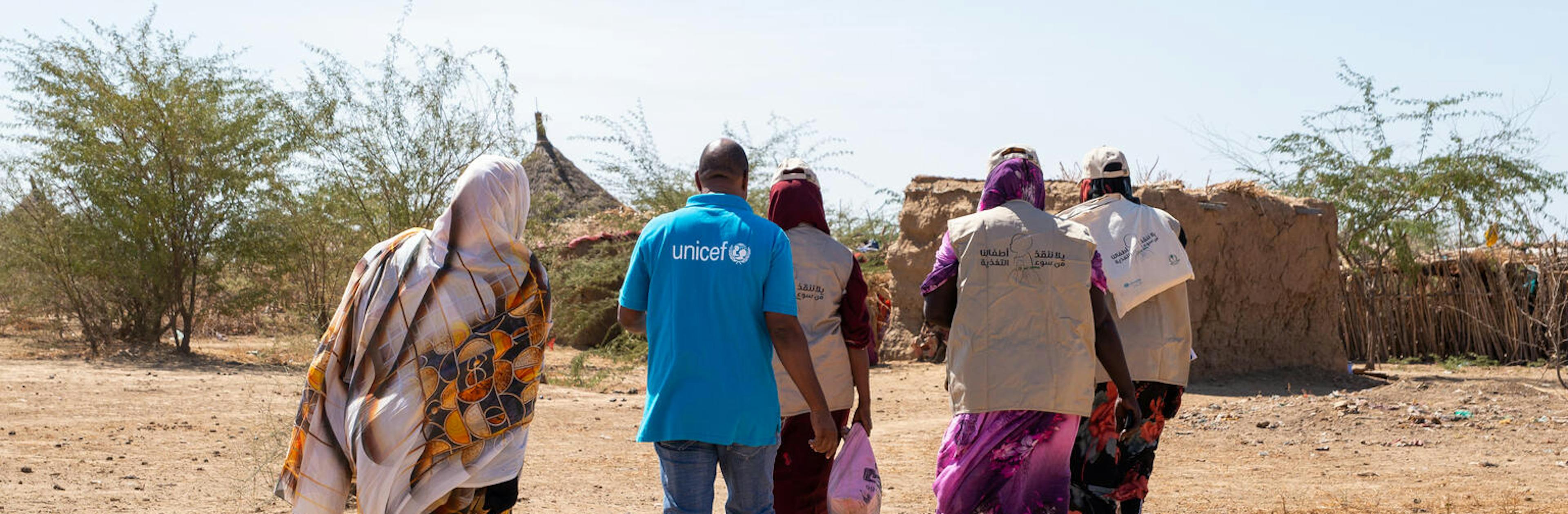 Aid workers wearing UNICEF uniforms walk through a rural area toward a dwelling.