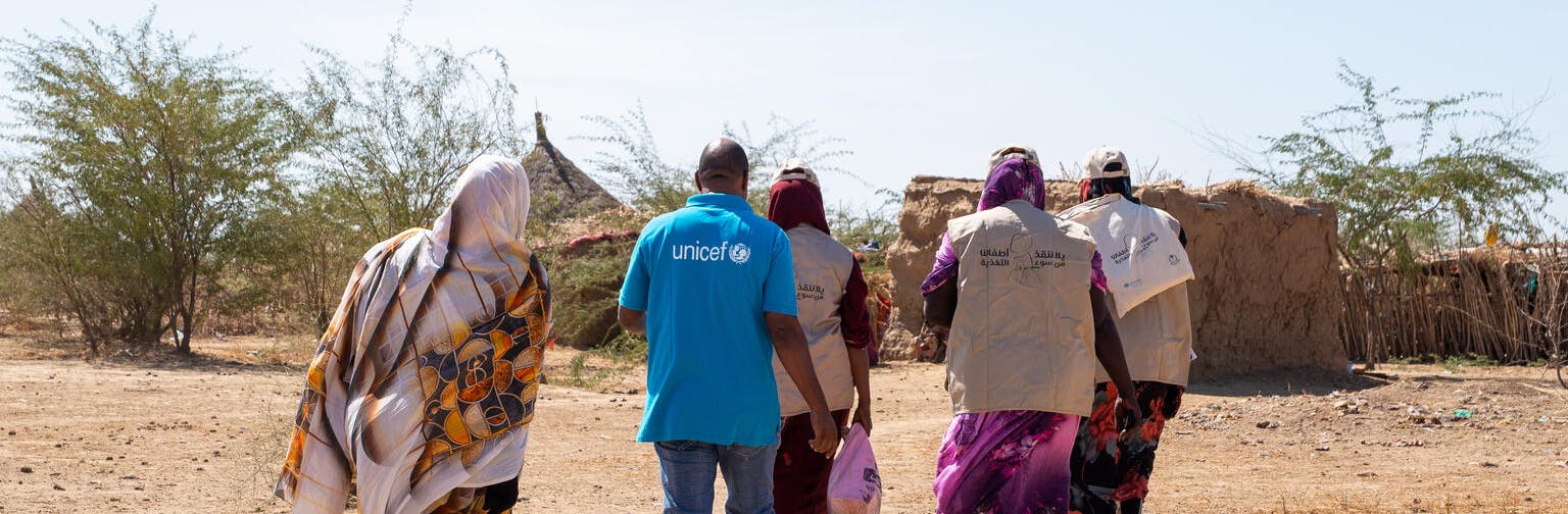 Aid workers wearing UNICEF uniforms walk through a rural area toward a dwelling.