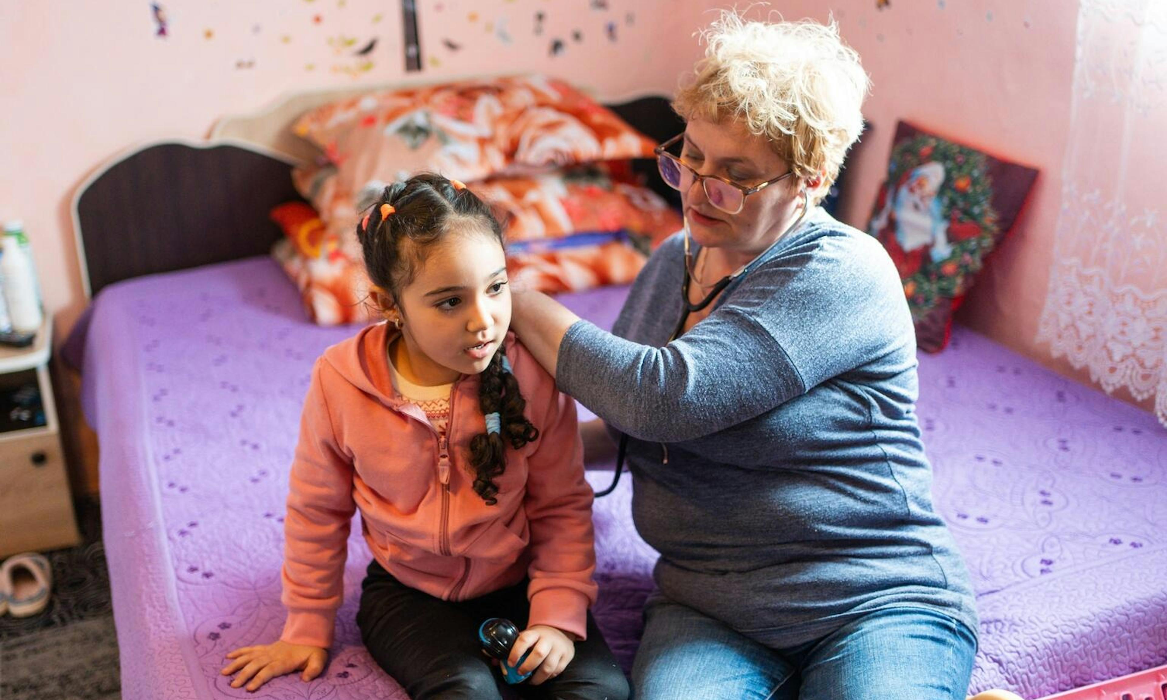 young girl sits on bed while women listens to her heart through a stethoscope