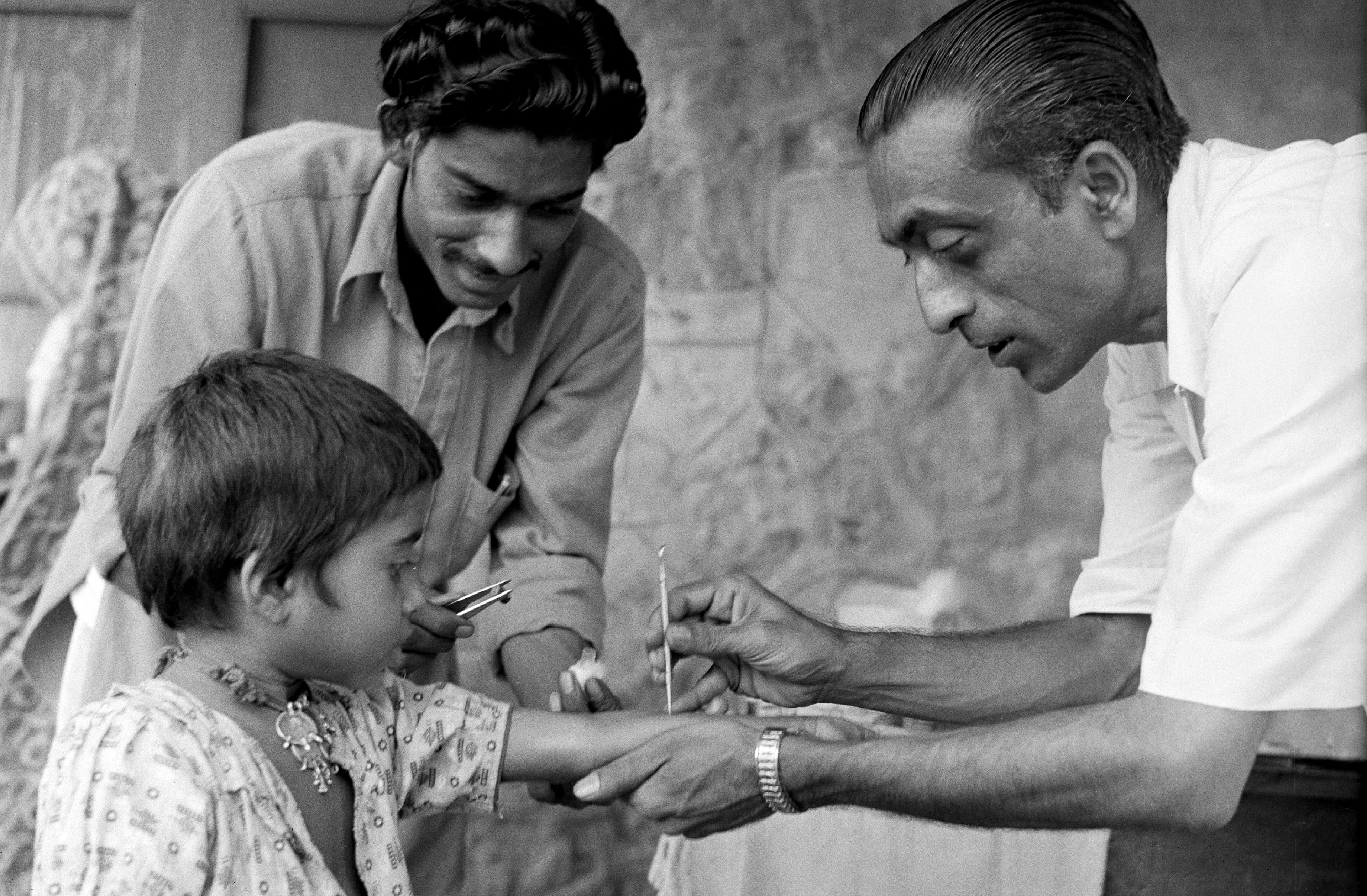 A girl watches a man health worker vaccinate her against smallpox.