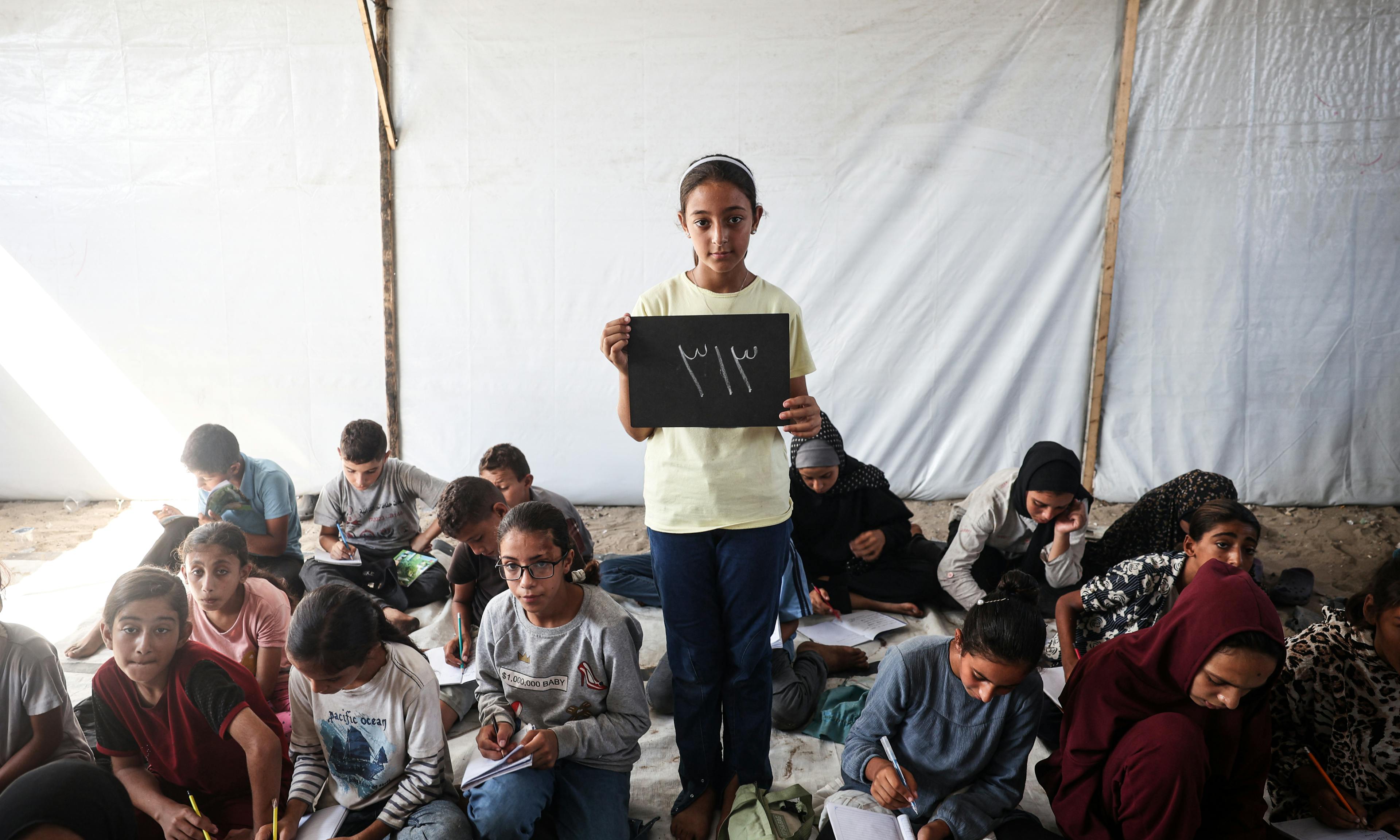 At the Deir Al-Balah Camp in the Gaza Strip, State of Palestine, 11-year-old Elham holds a chalkboard showing the number of school days she has missed, 313.