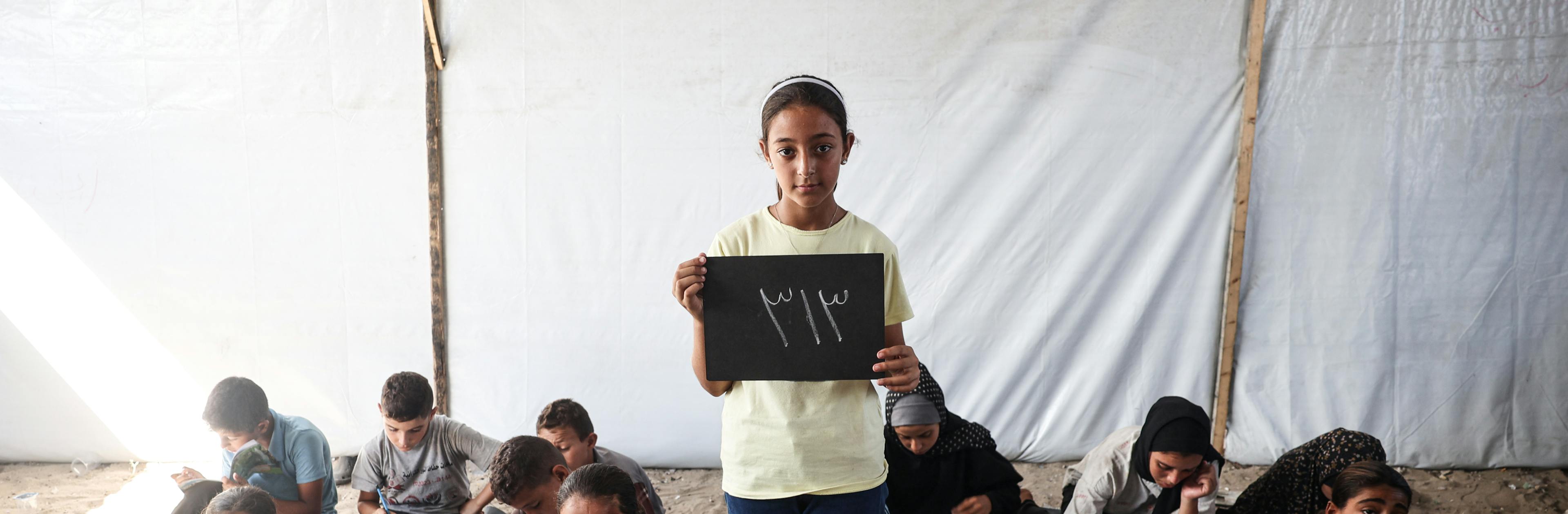 At the Deir Al-Balah Camp in the Gaza Strip, State of Palestine, 11-year-old Elham holds a chalkboard showing the number of school days she has missed, 313.