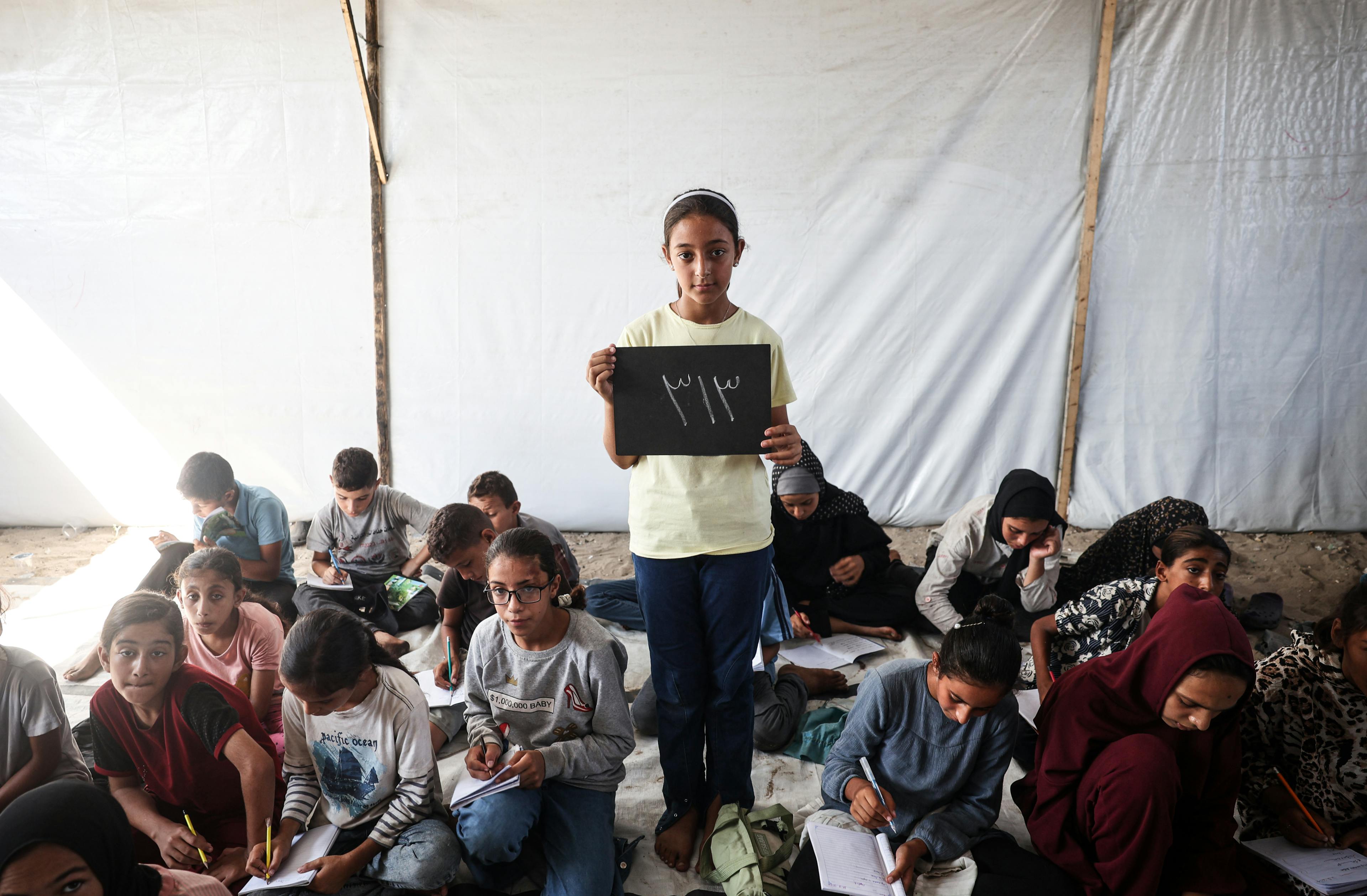 At the Deir Al-Balah Camp in the Gaza Strip, State of Palestine, 11-year-old Elham holds a chalkboard showing the number of school days she has missed, 313.