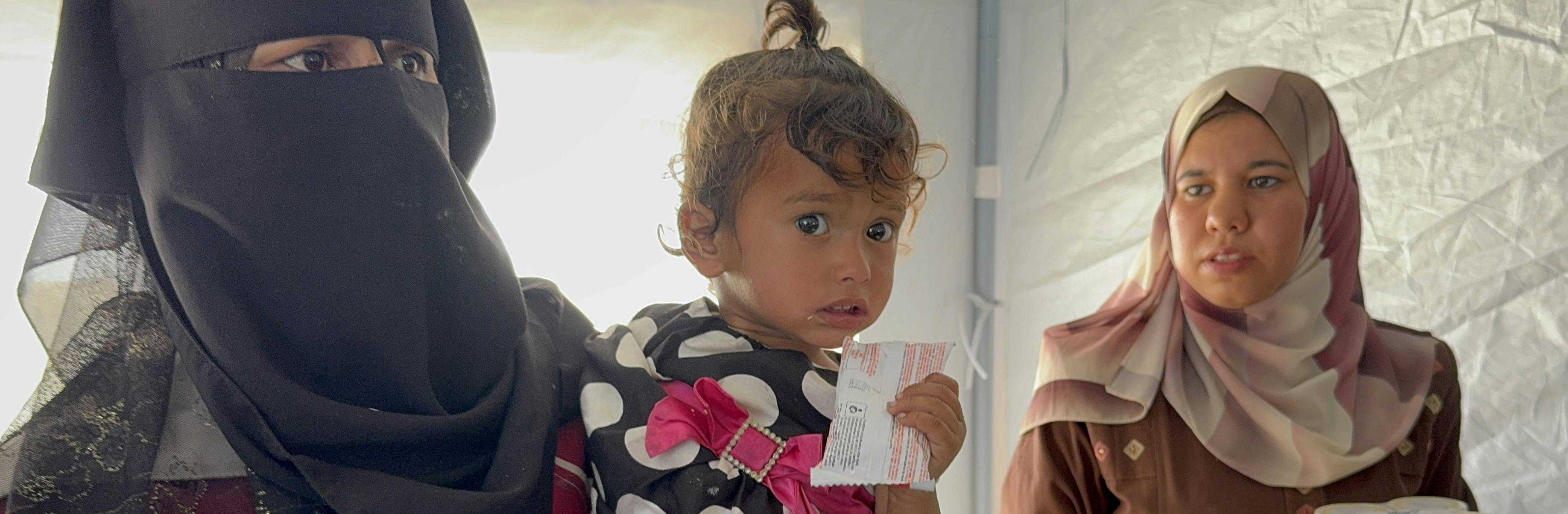 at Al Farooq Camp in Khan Yunis, a woman holds a small child who grips a sachet of Ready-to-Use Therapeutic Food (RUTF). In Al Farooq Camp, Khan Yunis, UNICEF has established a dedicated clinic to identify and treat children suffering from acute malnutrition. For over two months, the blockade has prevented vital medical and nutrition supplies from entering the Gaza Strip, leaving an estimated 5,000–6,000 children in need of therapeutic care each month. At this clinic, trained staff conduct MUAC (mid-upper-arm circumference) screenings and distribute Ready-to-Use Therapeutic Food (RUTF) to help restore children’s health and build their resilience.