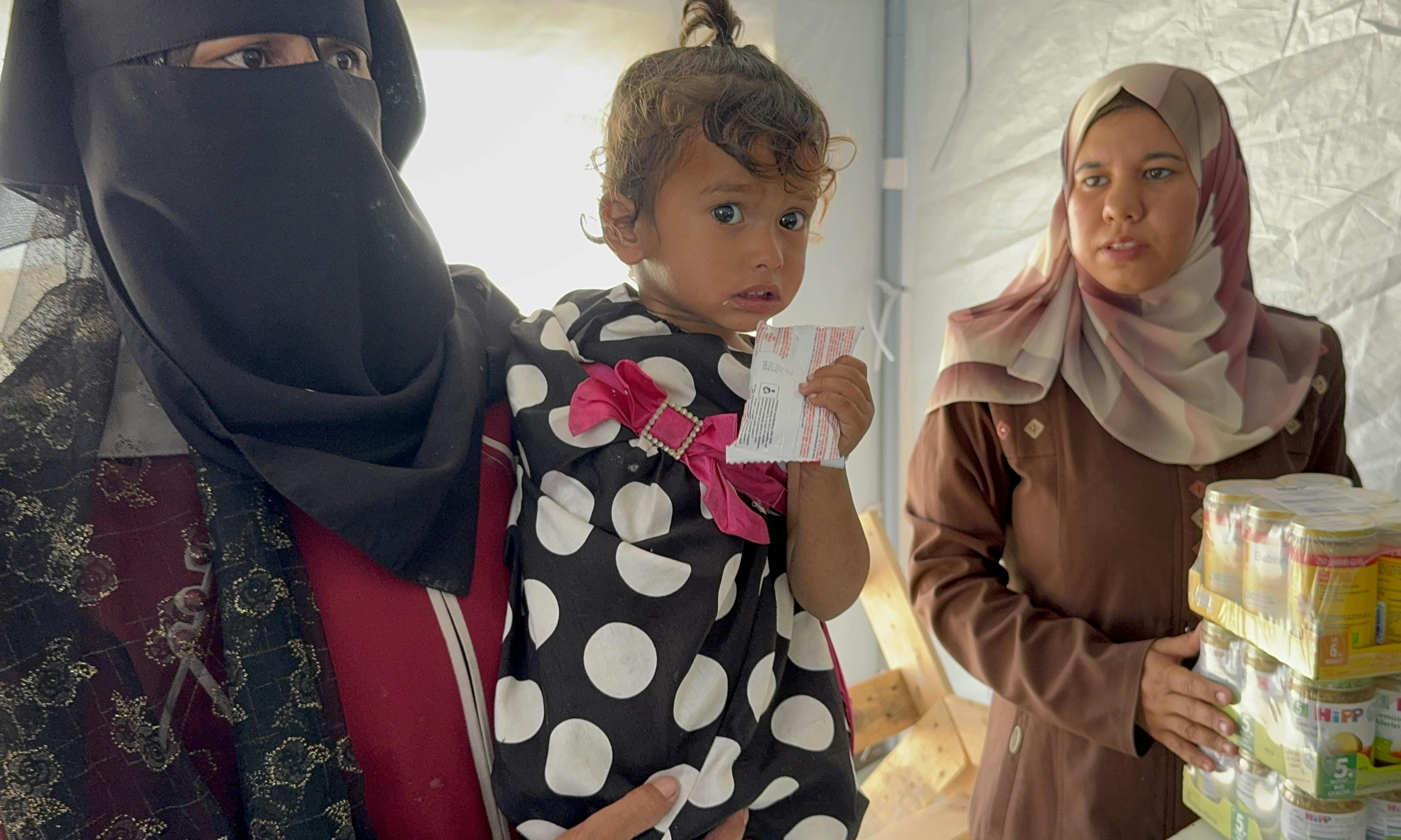 at Al Farooq Camp in Khan Yunis, a woman holds a small child who grips a sachet of Ready-to-Use Therapeutic Food (RUTF). In Al Farooq Camp, Khan Yunis, UNICEF has established a dedicated clinic to identify and treat children suffering from acute malnutrition. For over two months, the blockade has prevented vital medical and nutrition supplies from entering the Gaza Strip, leaving an estimated 5,000–6,000 children in need of therapeutic care each month. At this clinic, trained staff conduct MUAC (mid-upper-arm circumference) screenings and distribute Ready-to-Use Therapeutic Food (RUTF) to help restore children’s health and build their resilience.