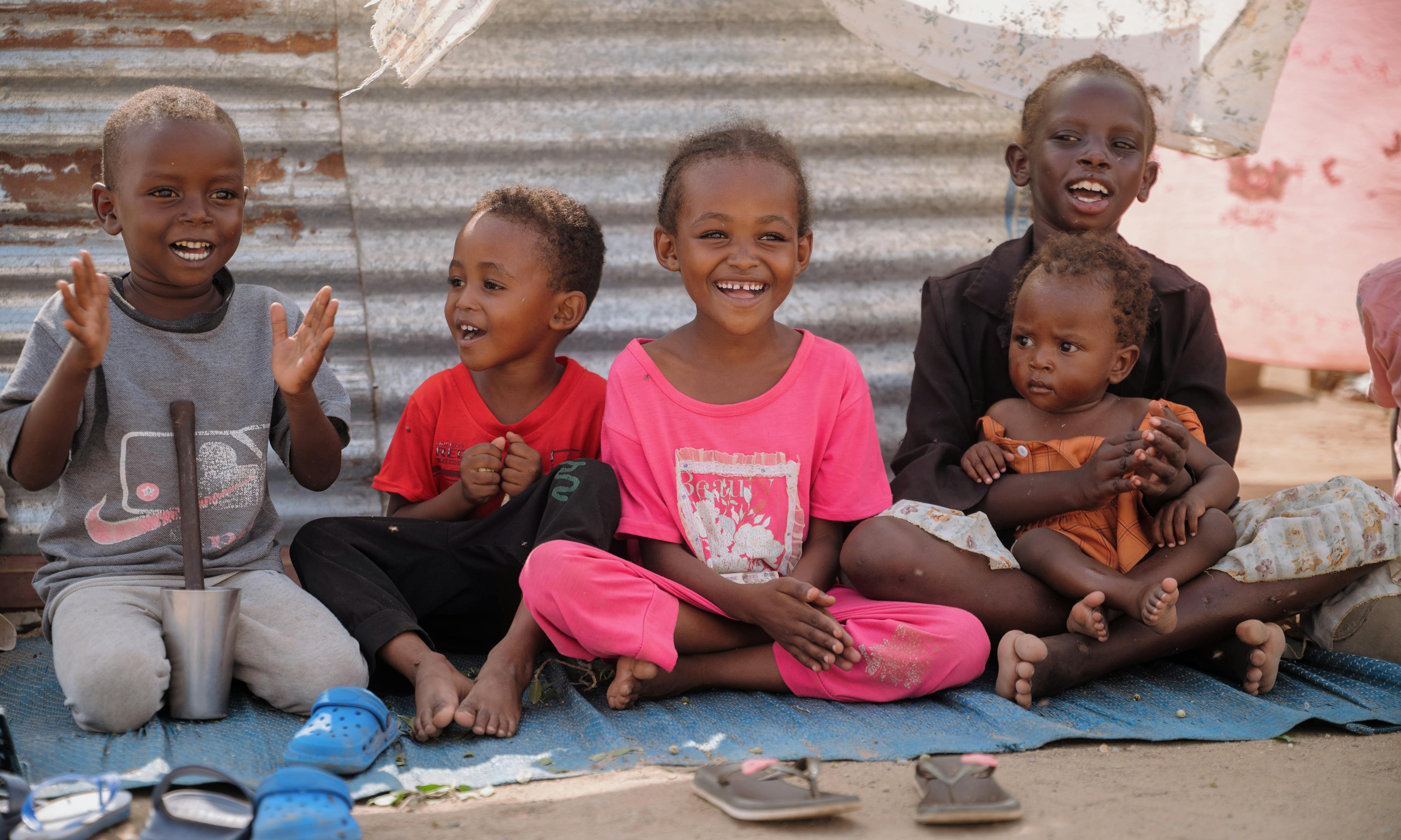 On 8 December 2025, children sit together outside their shelter at As-Senniya IDP site in Port Sudan, smiling and clapping as they enjoy a moment of play after fleeing violence across Sudan.