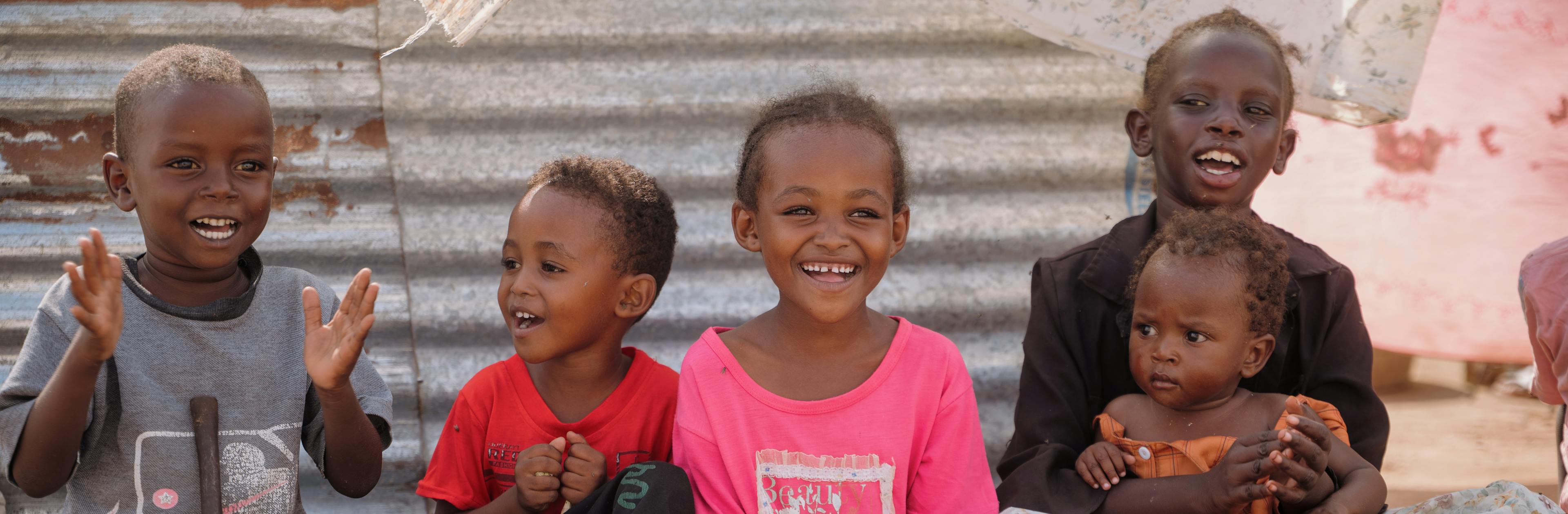 On 8 December 2025, children sit together outside their shelter at As-Senniya IDP site in Port Sudan, smiling and clapping as they enjoy a moment of play after fleeing violence across Sudan.