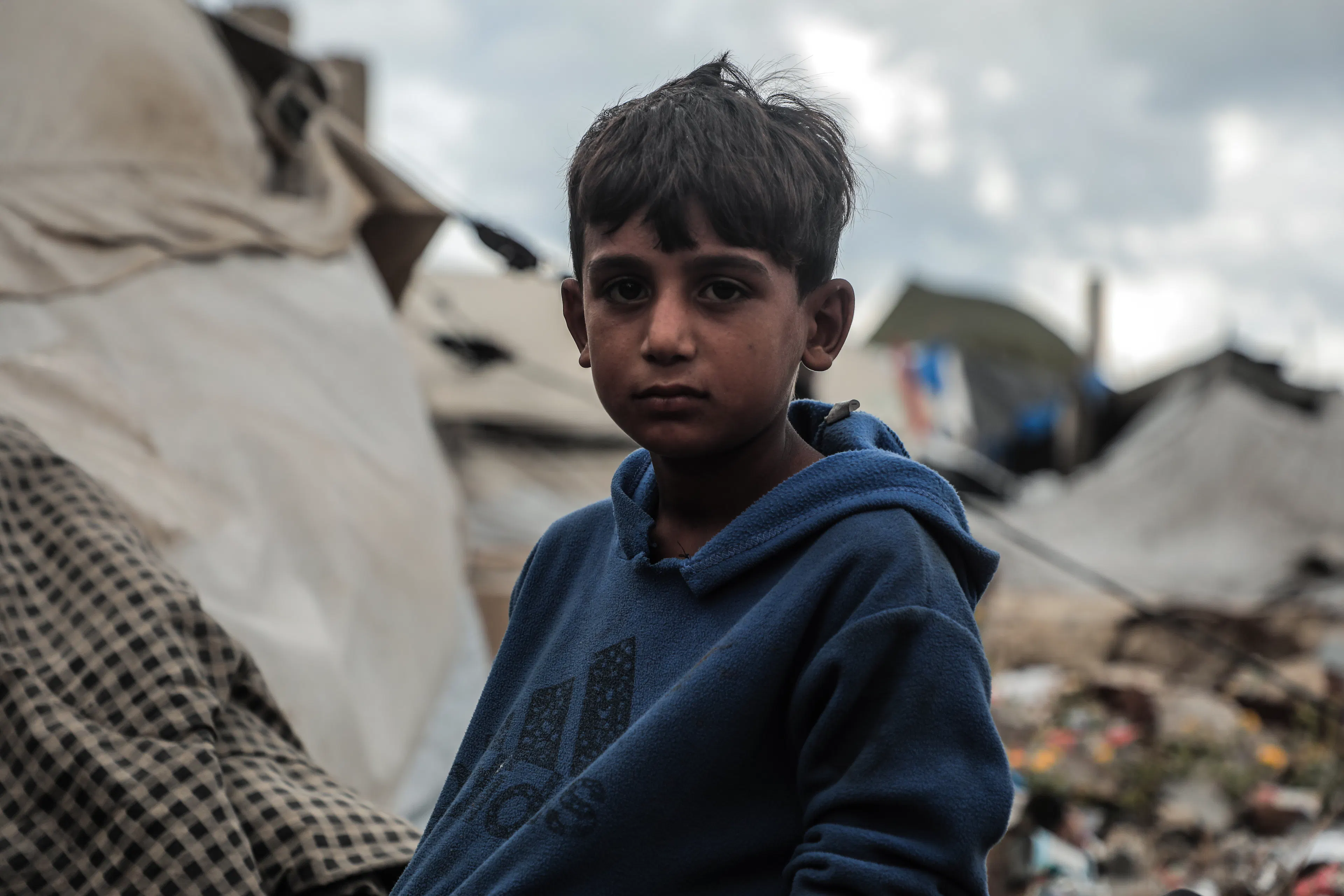 Boy wearing blue jumper in Gaza stands in front of rubble surrounded by tents
