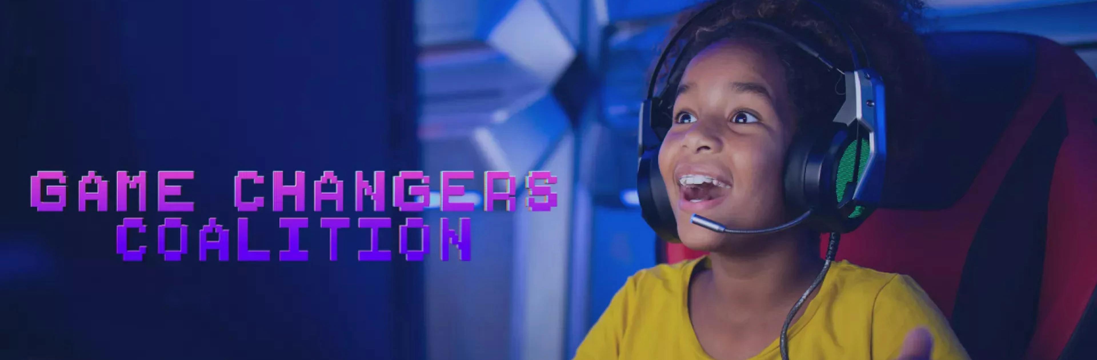 A young girl wearing a yellow shirt and a headset plays a video game on a computer with an expression of joy on her face in a darkly-lit room with purple lights.