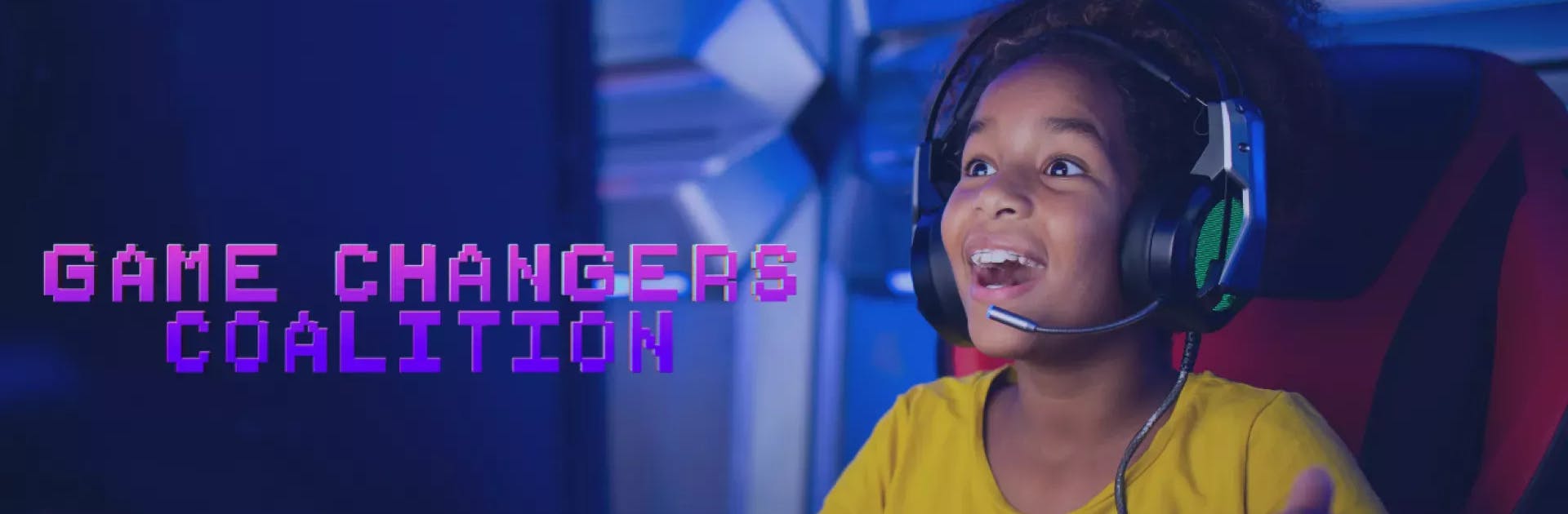 A young girl wearing a yellow shirt and a headset plays a video game on a computer with an expression of joy on her face in a darkly-lit room with purple lights.