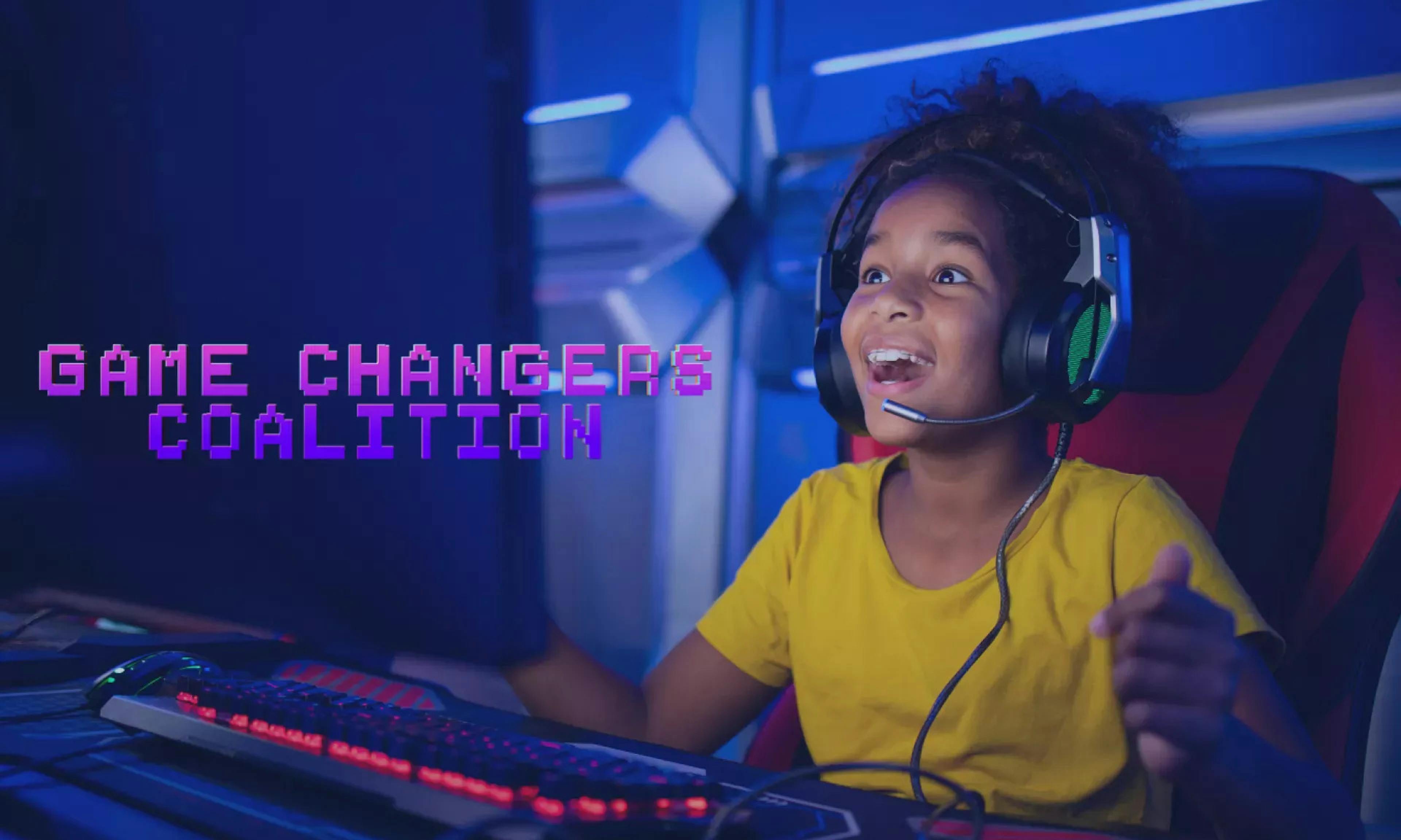 A young girl wearing a yellow shirt and a headset plays a video game on a computer with an expression of joy on her face in a darkly-lit room with purple lights.