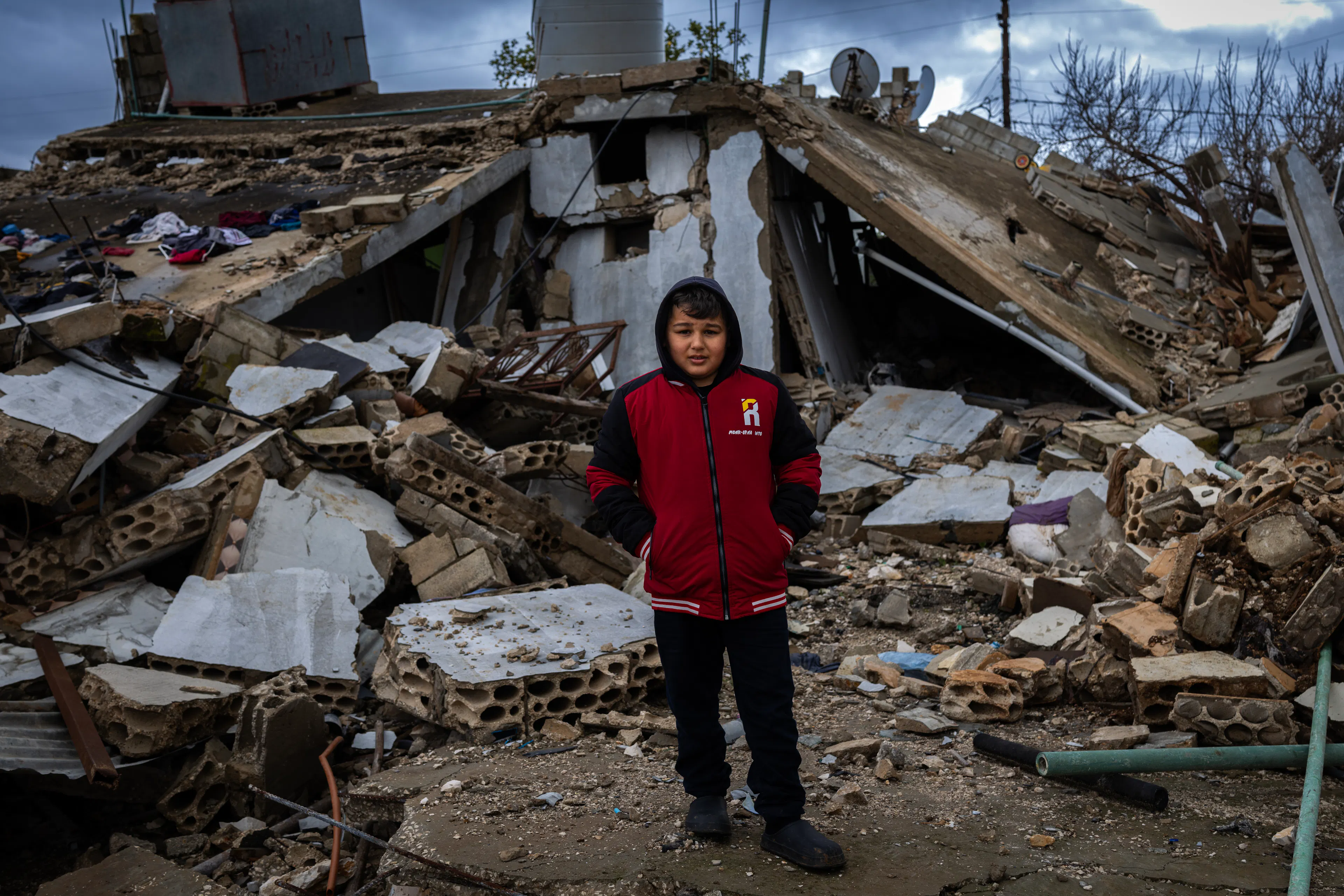 Boy wearing red jumper in Gaza stands in front of rubble
