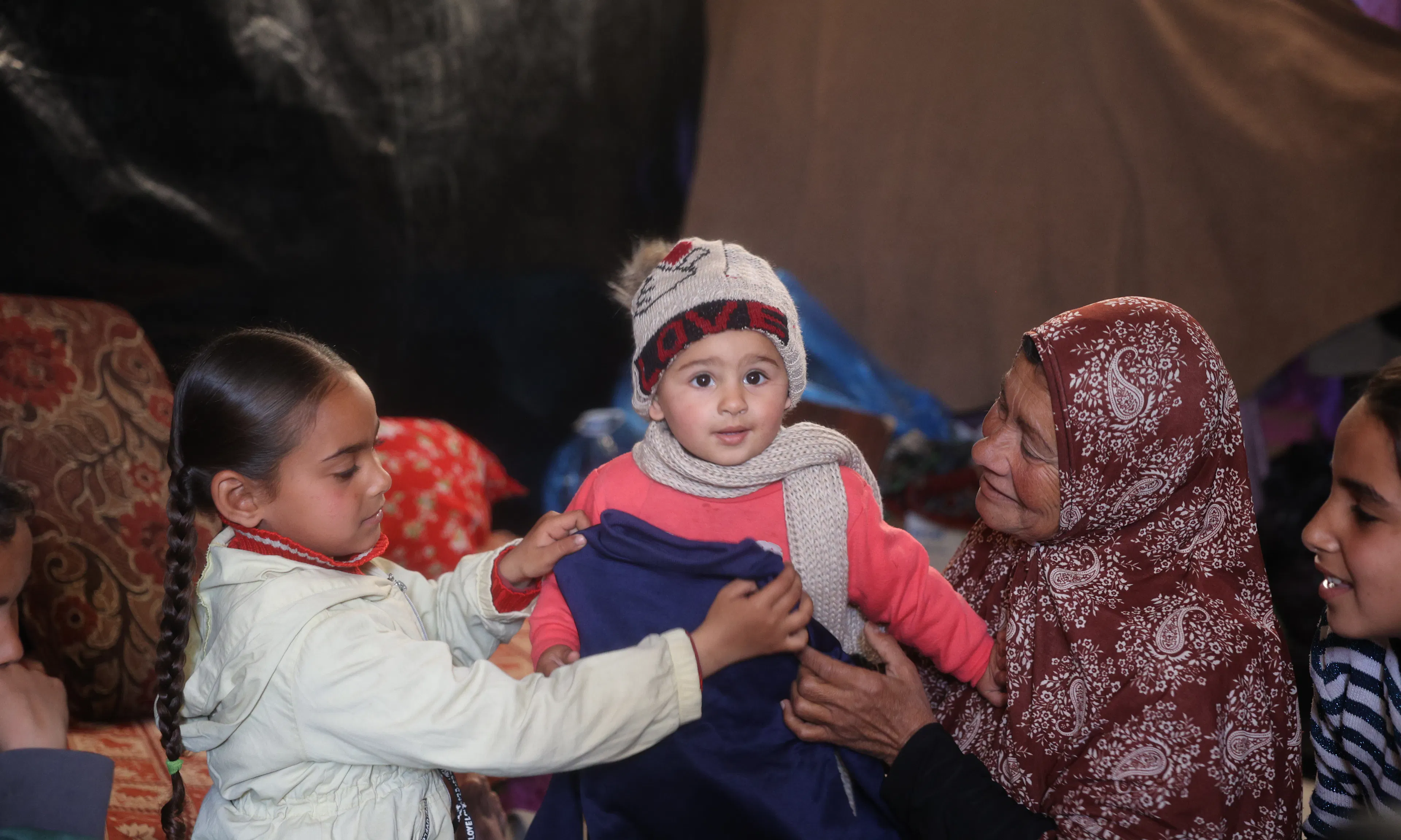 An elderly woman and a young girl smiling as they help a toddler put on a warm blue sweater and a knitted "LOVE" beanie in a sheltered living space.
