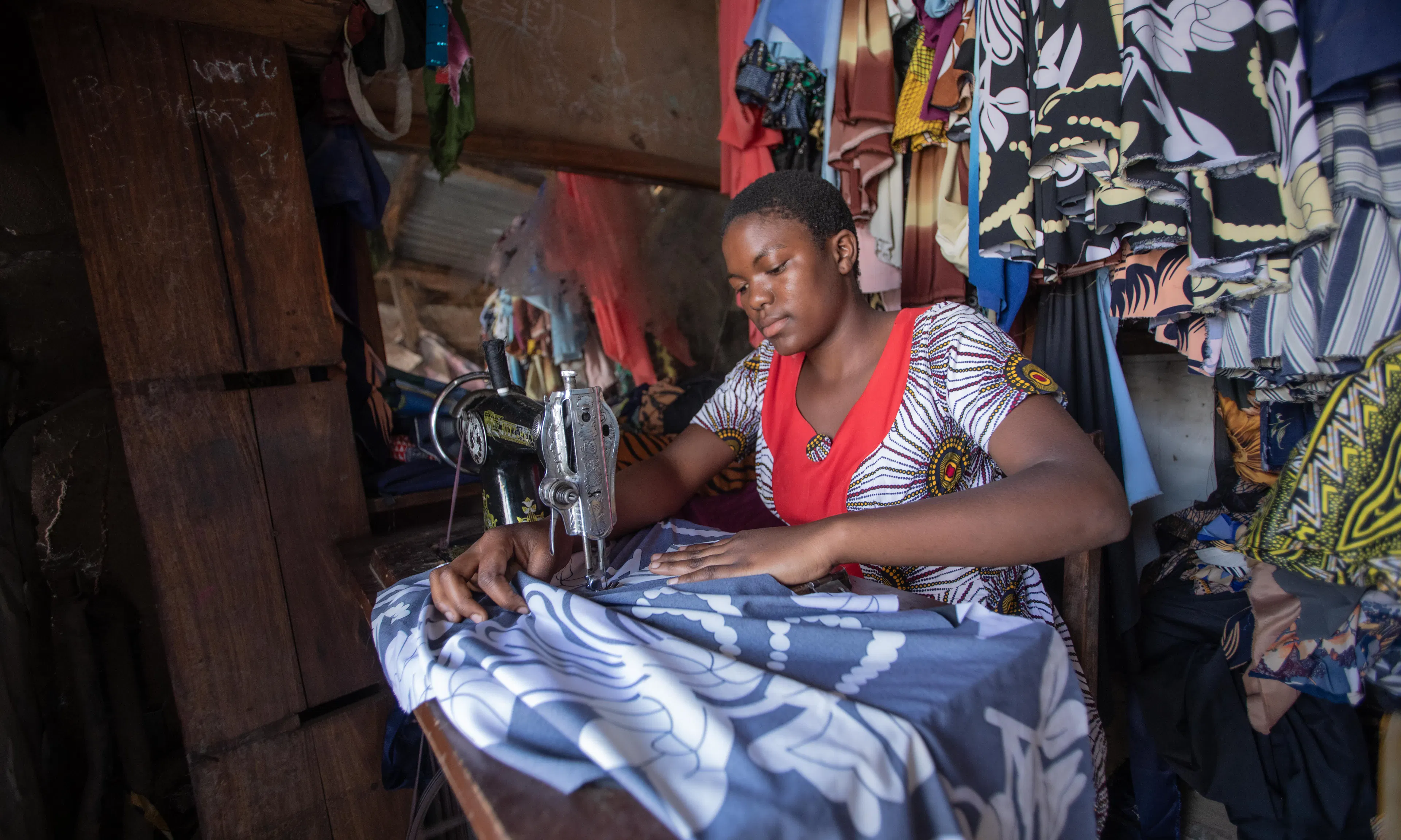 On 13 August 2025, Antini Mahepa, 17, works as a sewing apprentice in a local market located in the Uvinza District of the Kigoma Region in Tanzania. The small stall, lined with colourful fabrics and garments, is a place for learning and growth.
