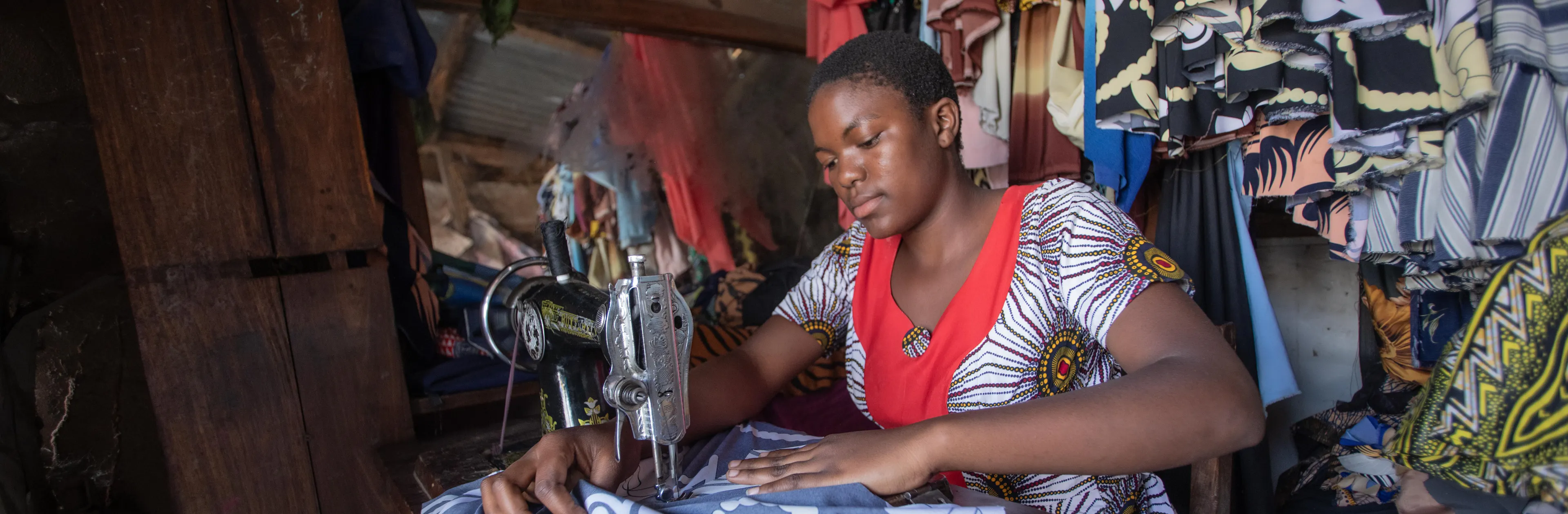On 13 August 2025, Antini Mahepa, 17, works as a sewing apprentice in a local market located in the Uvinza District of the Kigoma Region in Tanzania. The small stall, lined with colourful fabrics and garments, is a place for learning and growth.