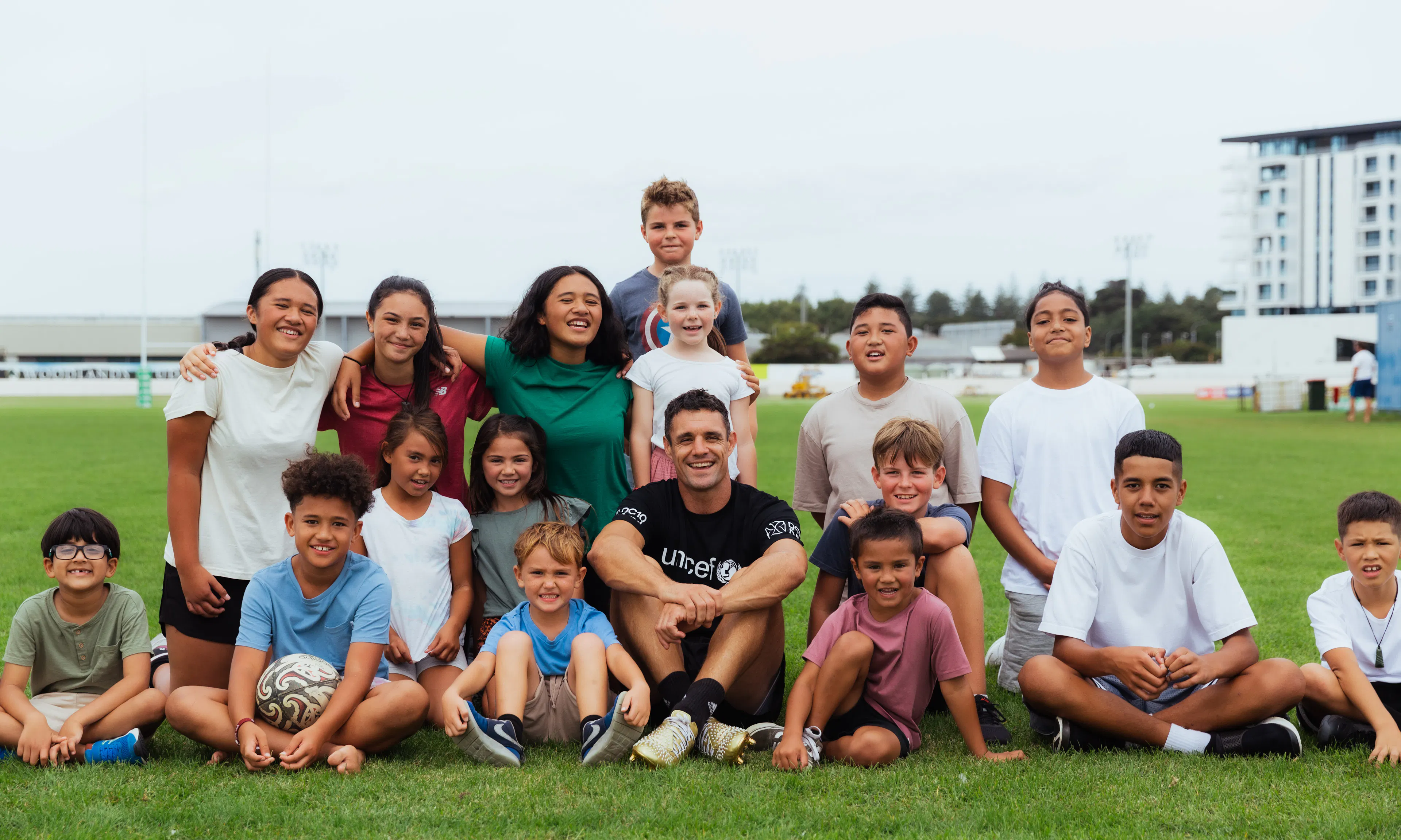 New Zealand rugby legend Dan Carter sits on a field with children before his Kickathon challenge to raise funds for providing clean water in the Pacific with UNICEF.
