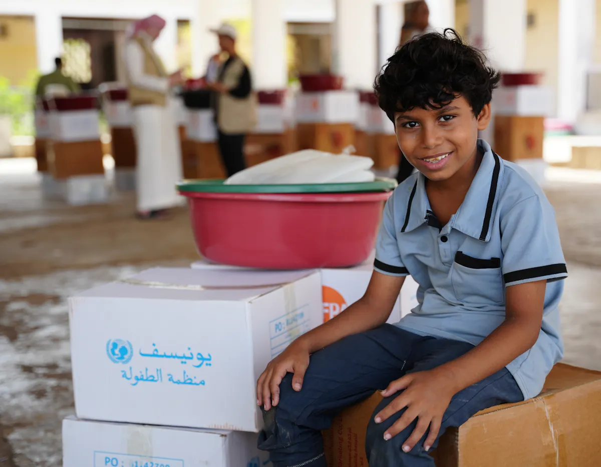 A child sits beside boxes of humanitarian supplies in Aden, Yemen. These supplies will improve health, prevent disease, and strengthen community resilience.