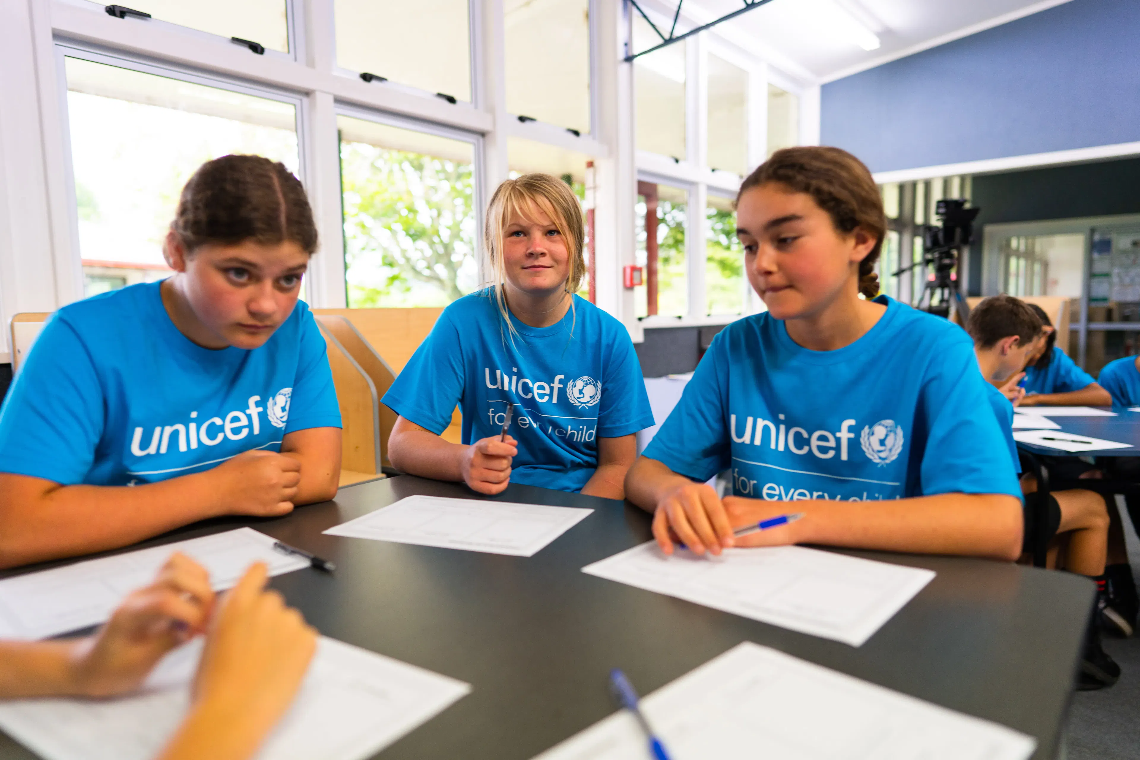 hree young girls in blue UNICEF shirts sitting around a desk at Poipoi School, participating in a group discussion with worksheets and pens.