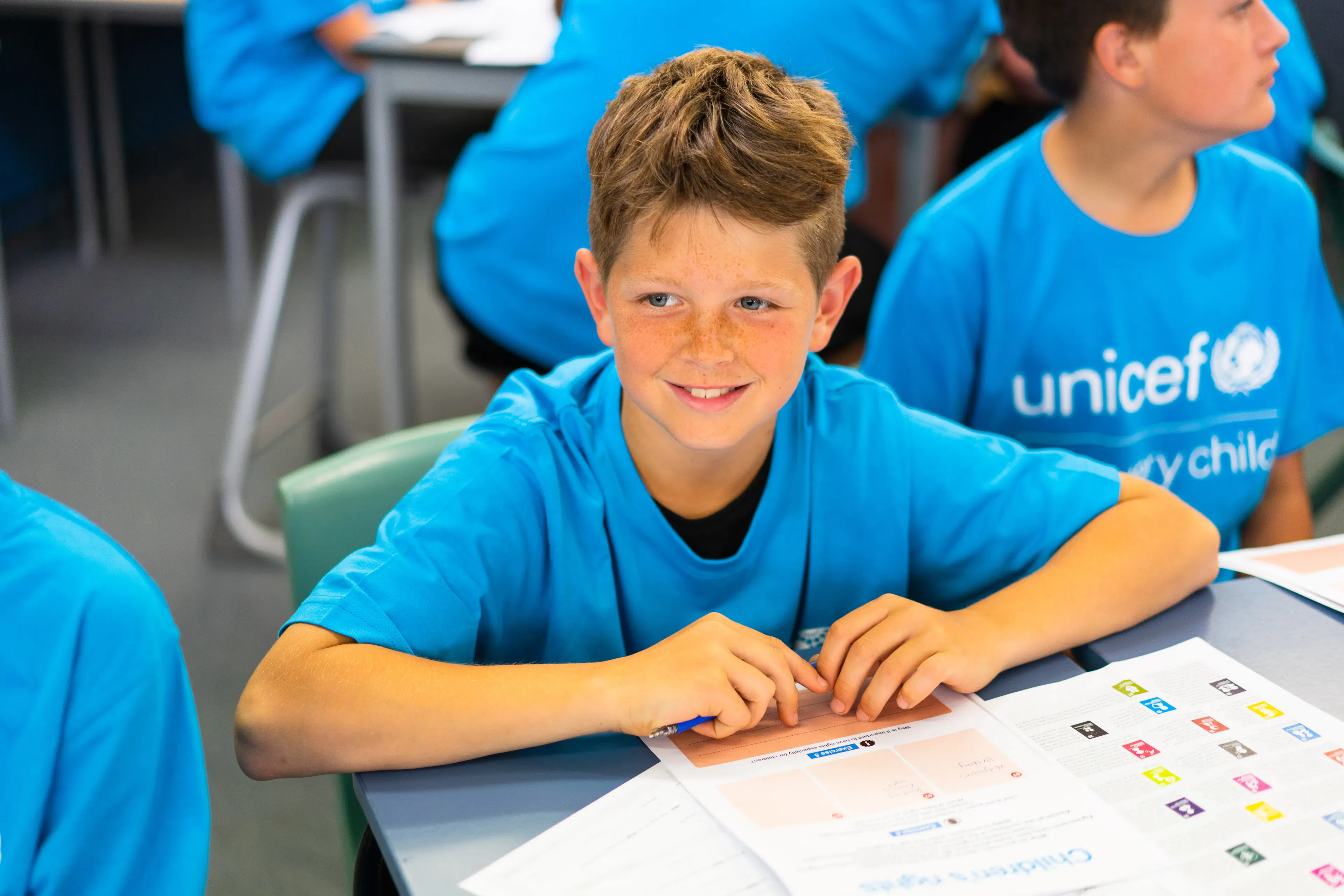 A young boy smiling and looking ahead while sitting at a desk with UNICEF educational worksheets during a school visit in November 2022.
