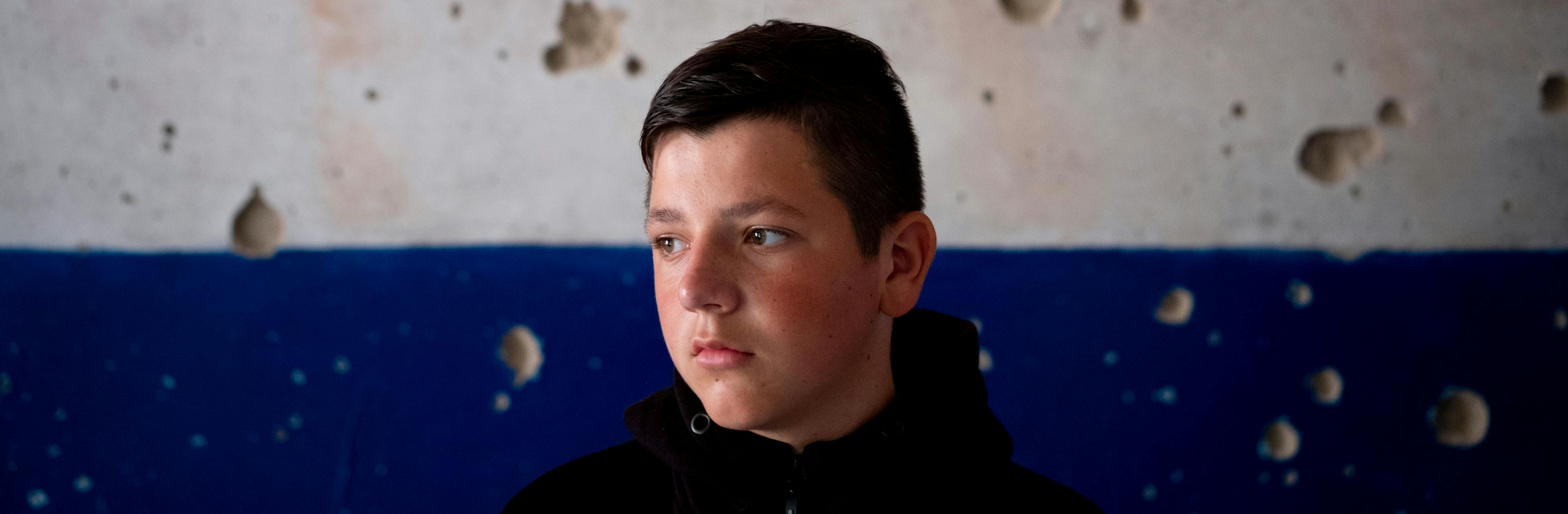 On 13 August 2022, Nazar, 13, stands in front of a scarred wall in his severely damaged school in the village of Olyzarivka, Ukraine.