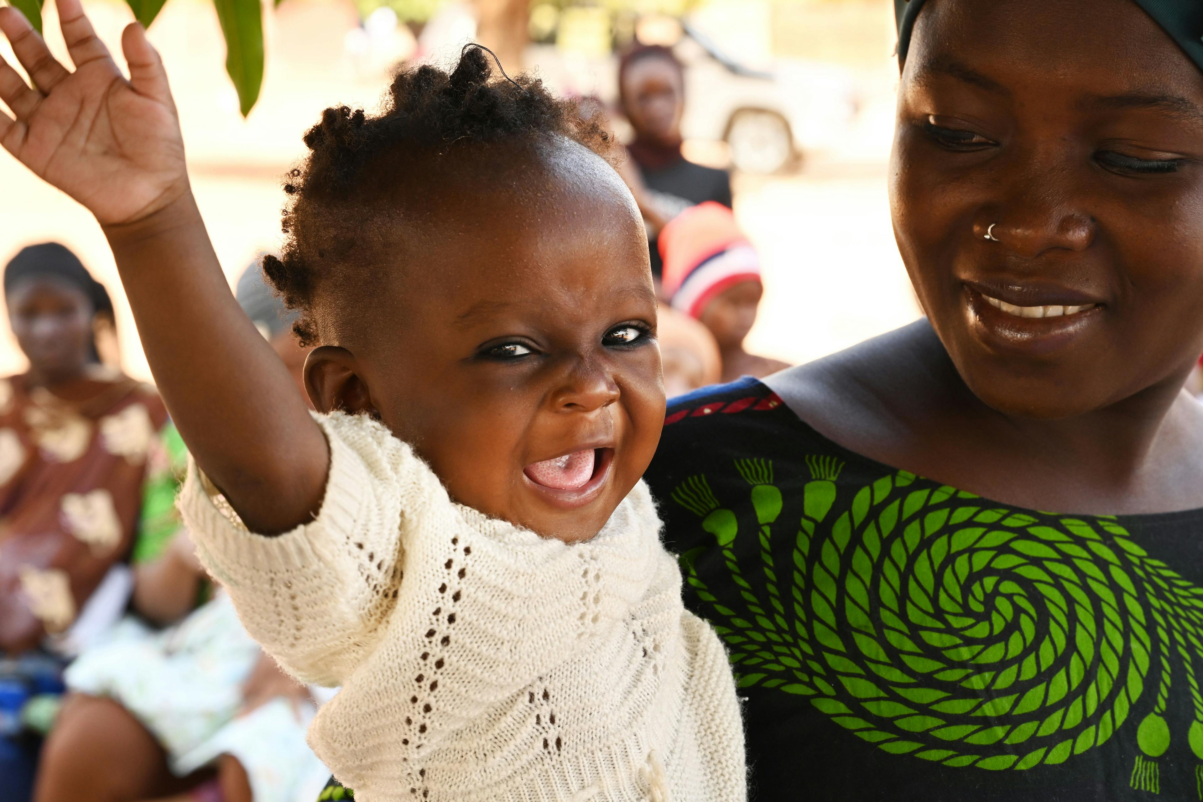 A toddler smiles and waves her hand while sitting in her mothers arms