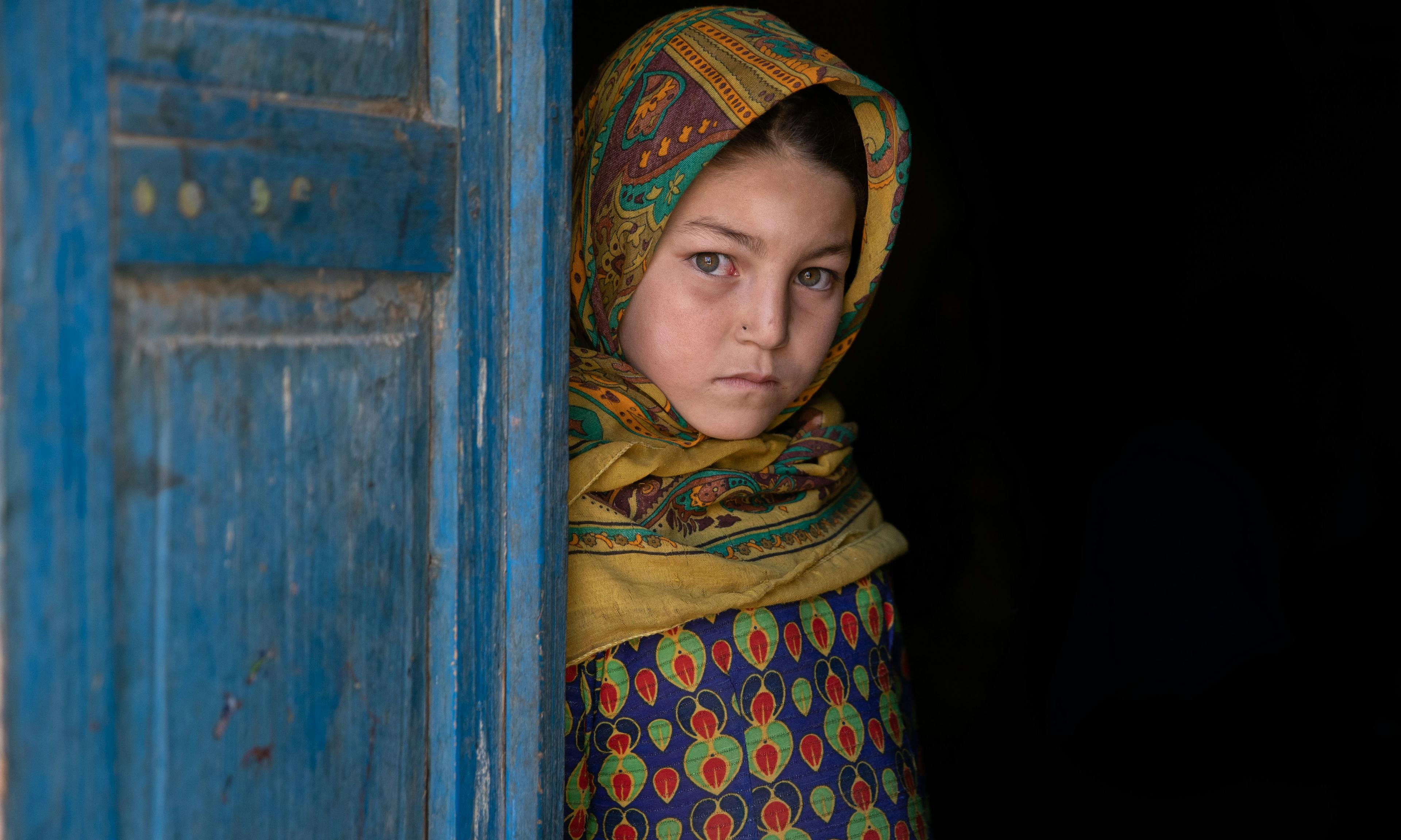 A sad girl peering behind a blue door in Afghanistan, symbolising the silent struggles of early marriages.