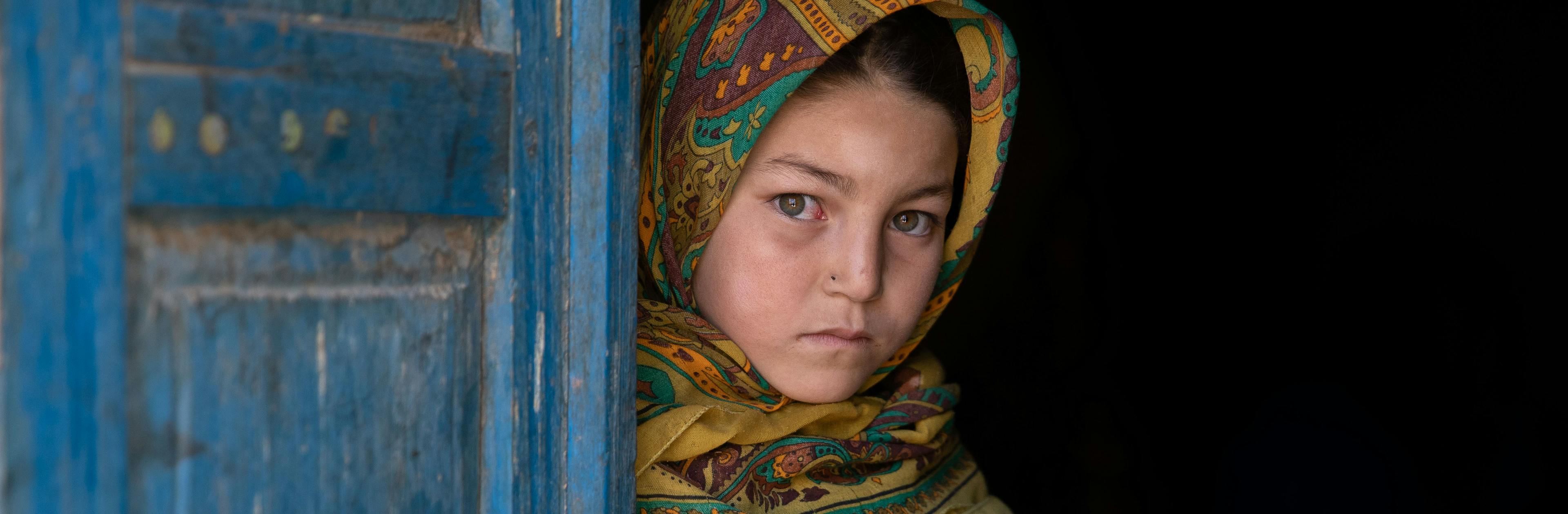 A sad girl peering behind a blue door in Afghanistan, symbolising the silent struggles of early marriages.