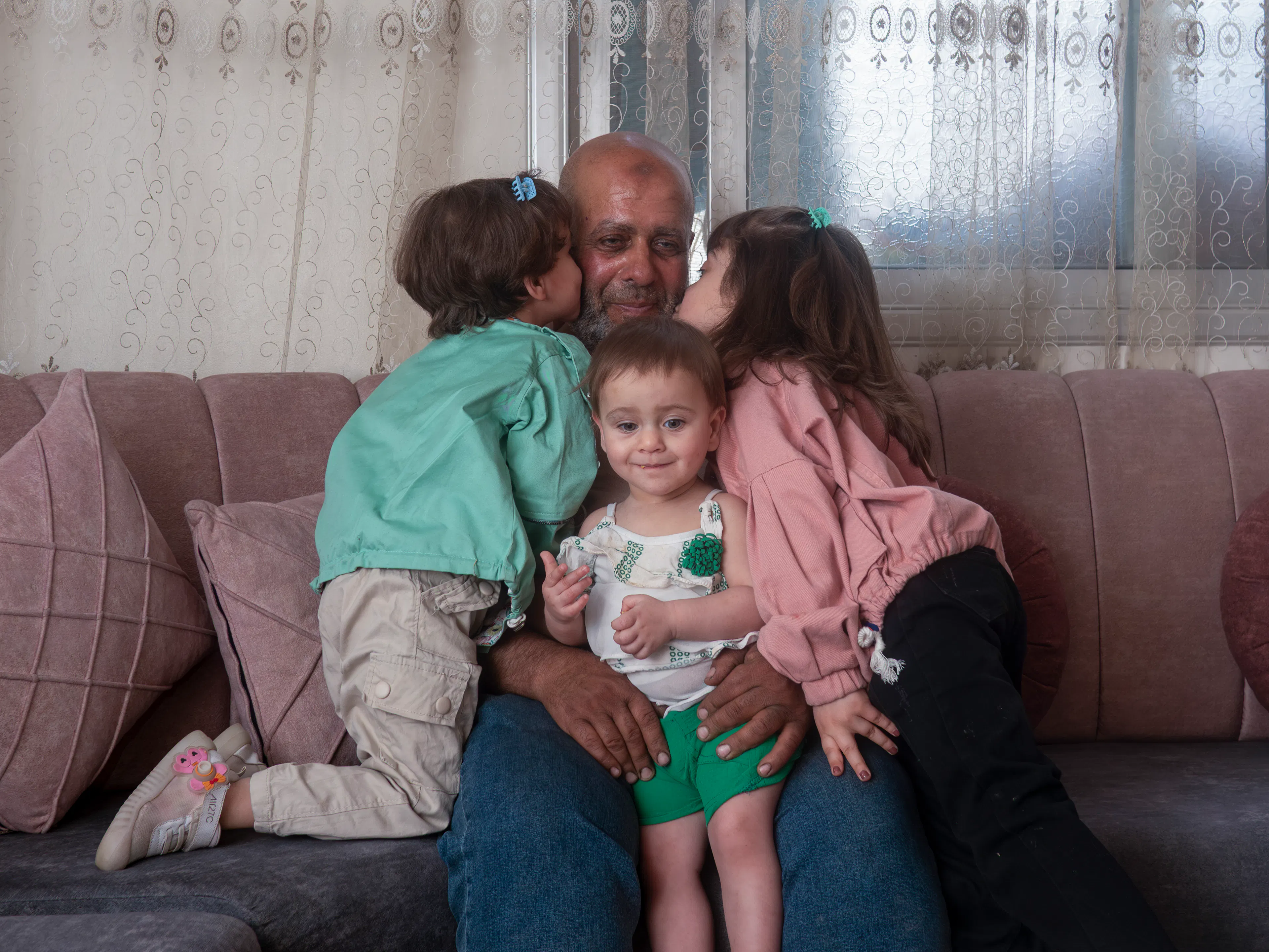 Aline, 5, and Lareen, 3, kiss their father, Abu Maher, 54, as he holds his youngest daughter Haneen at their home in Duma, Rural Damascus, Syria, on 19 May 2025.