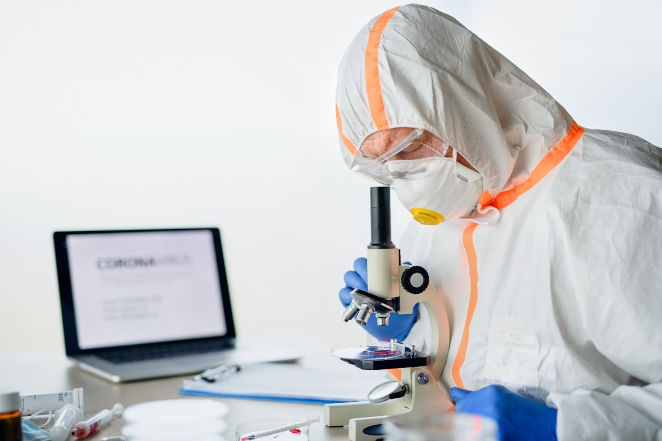 Lab technician inspecting something under a microscope with a computer running in the background and a desk with a clipboard and other equipment