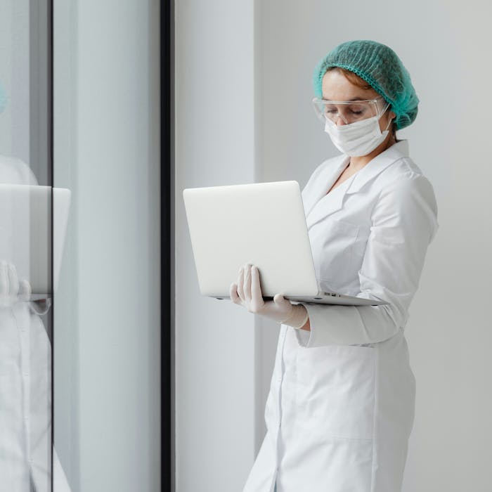 A scientist dressed in a white lab coat examines data on a laptop screen that she is carrying