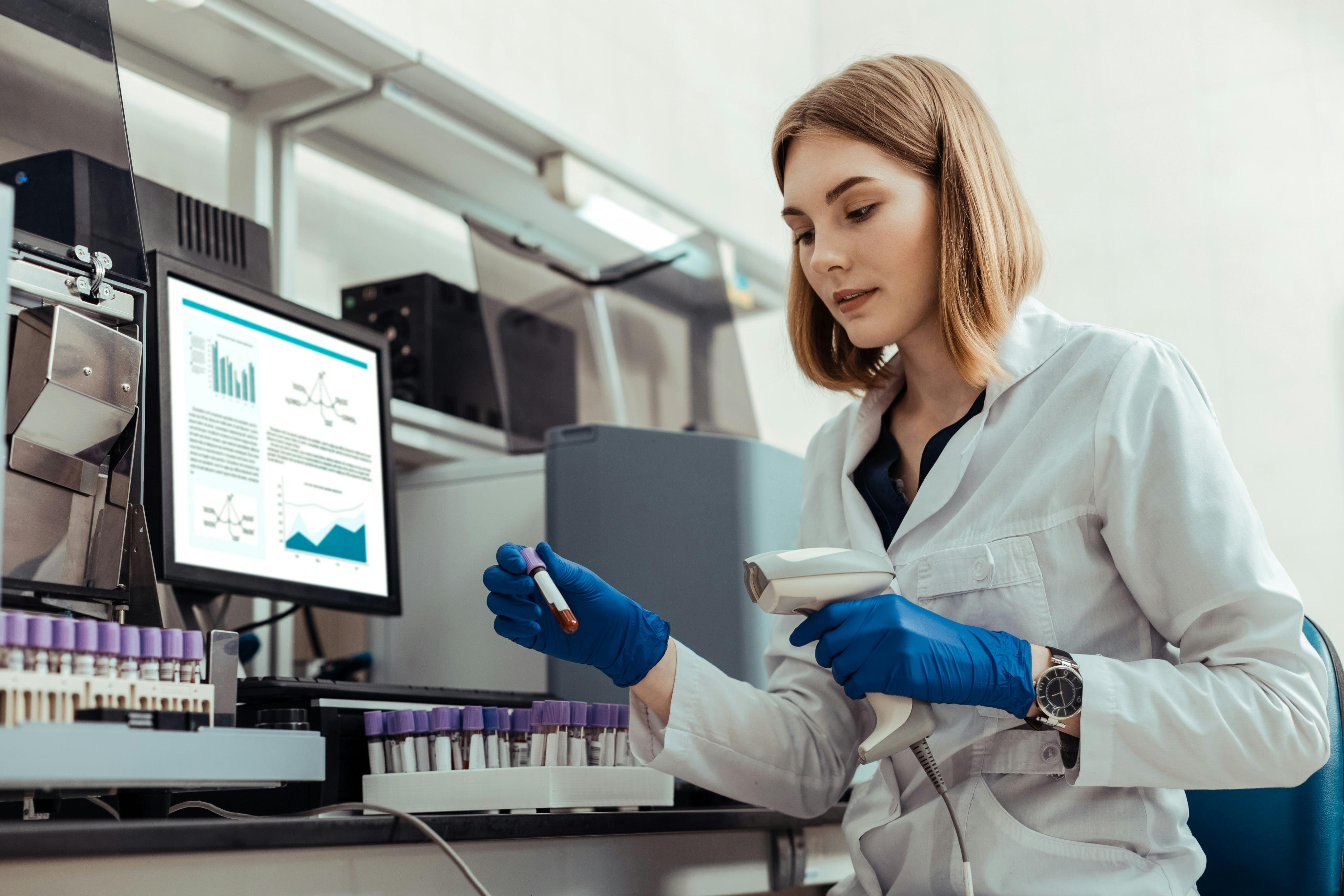 A scientist in a lab conducts an experiment with a test tube and a hand-held scanning device, while a computer in the background shows a dashboard of data