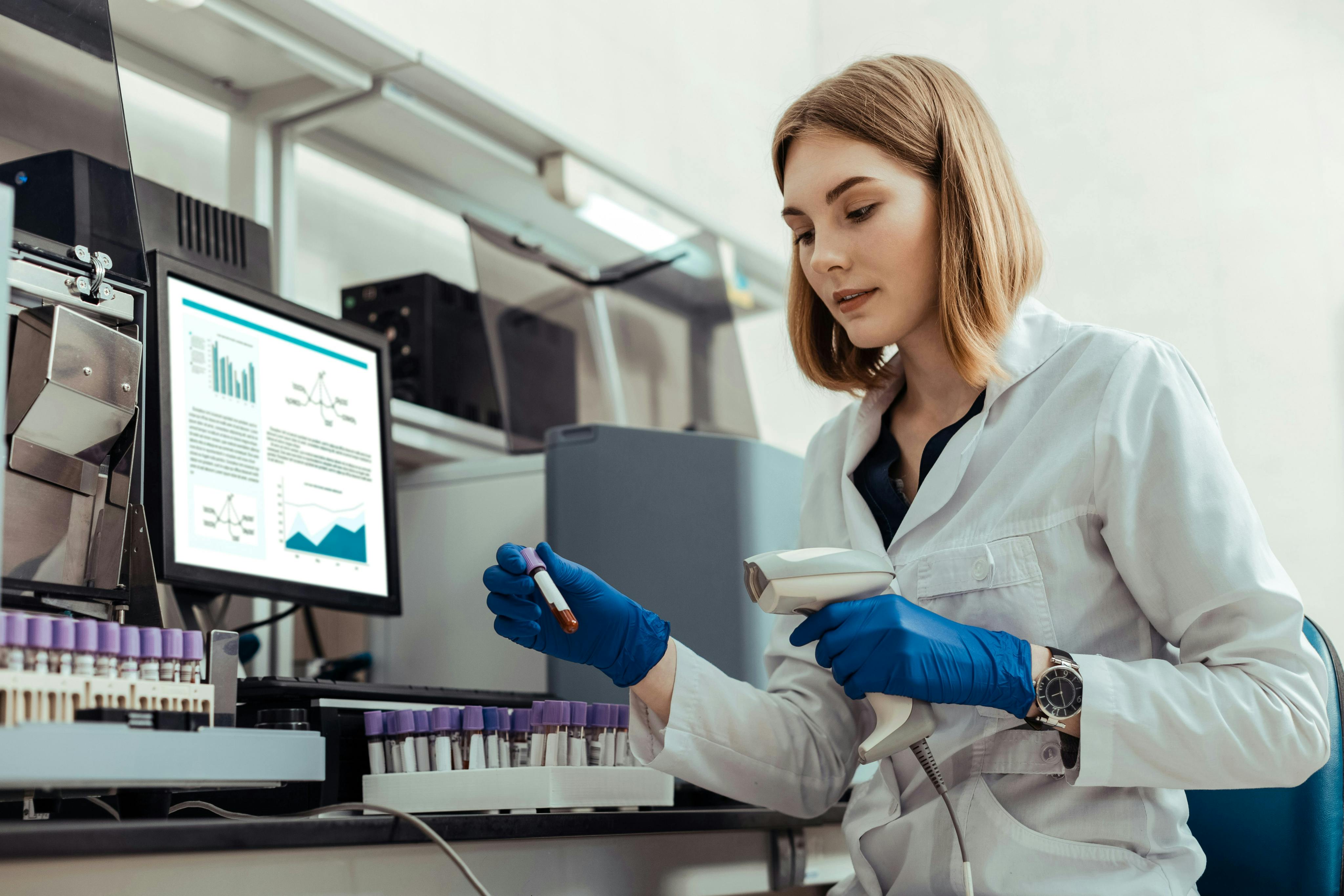 A scientist in a lab conducts an experiment with a test tube and a hand-held scanning device, while a computer in the background shows a dashboard of data