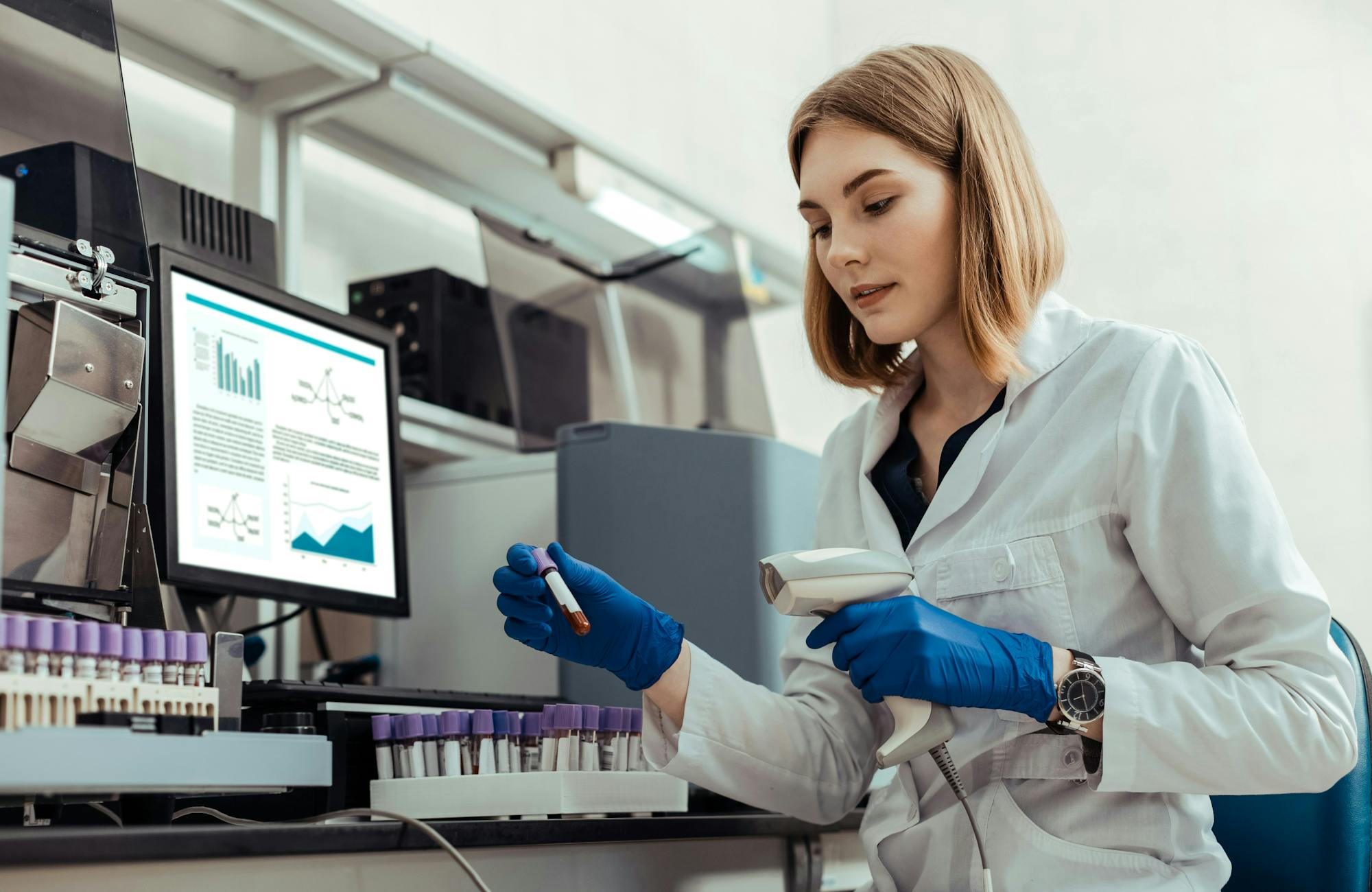 A scientist in a lab conducts an experiment with a test tube and a hand-held scanning device, while a computer in the background shows a dashboard of data