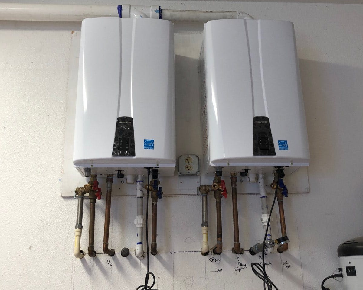 Two white wall-mounted water heaters side by side, each with a digital control panel. Below each heater are various pipes and valves connected. Annotations and measurements are scribbled on the wall beneath them