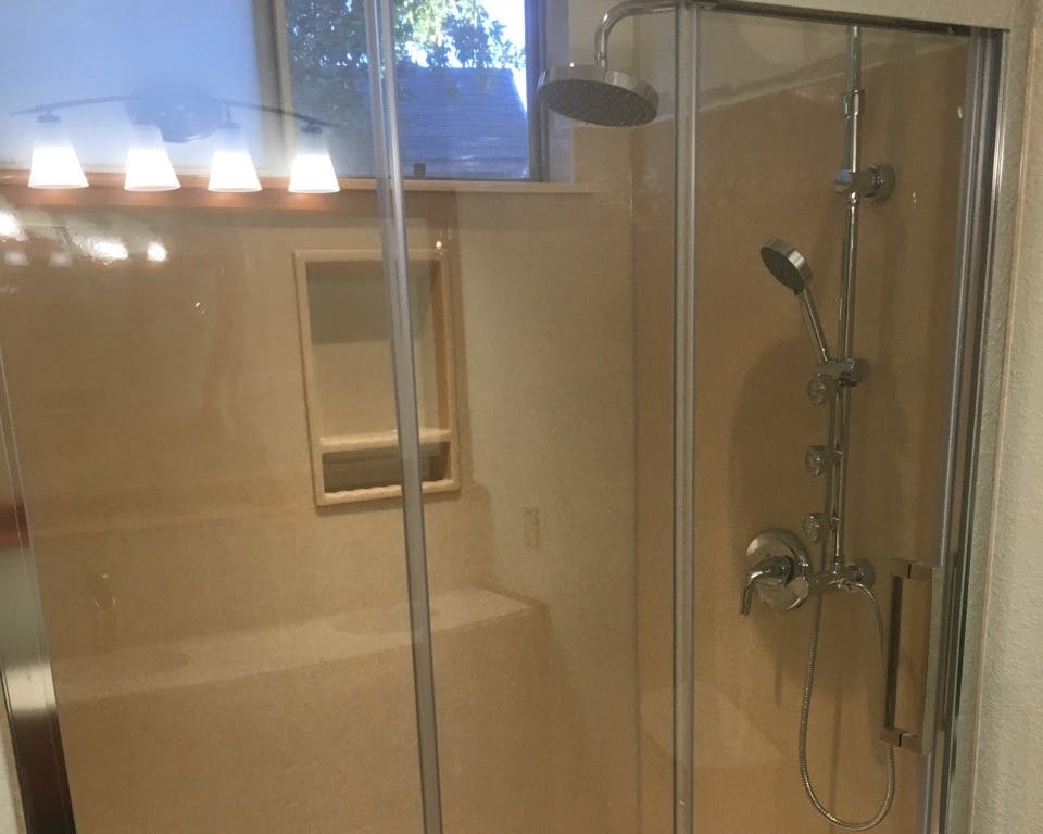 A clear glass shower enclosure with silver fixtures. Inside, there's a dual showerhead system with both a rainfall and handheld option. The beige wall features a recessed shelf, and overhead lights are reflected at the top of the image. A window is partially visible to the left.