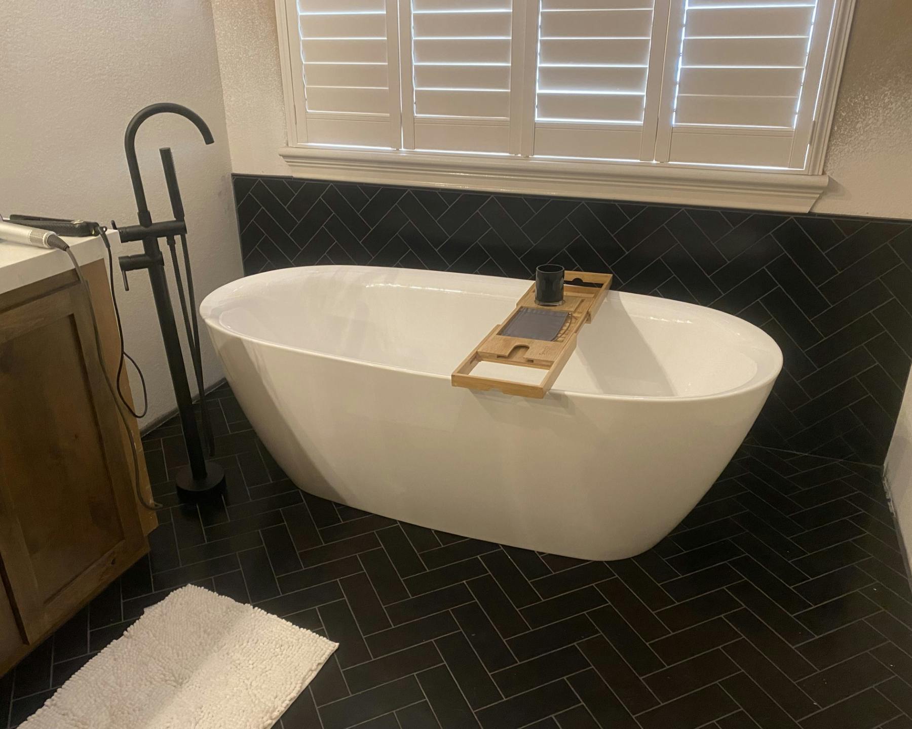"A contemporary bathroom showcasing a white freestanding bathtub beside a window with white horizontal blinds. On the tub, a wooden bath tray holds a dark mug. Adjacent to the tub is a dark floor-standing faucet. The lower half of the walls features black tiles with a diagonal design, complemented by a white wooden cabinet on the left. A soft white rug is spread on the floor.