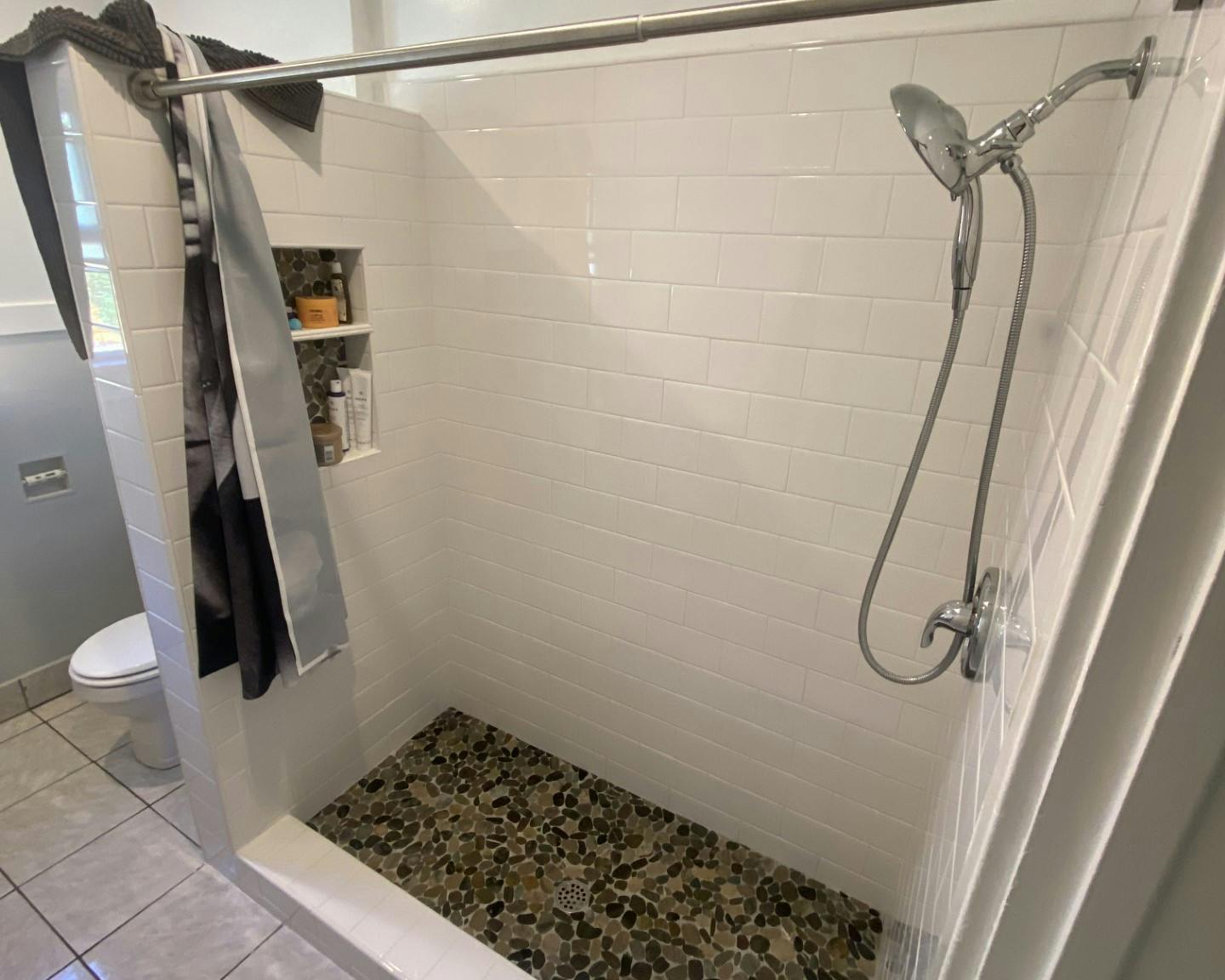 A well-lit bathroom with a walk-in shower showcasing white horizontal tiling on the walls and a pebble stone floor. A stainless steel handheld showerhead hangs from the right side. The left shows a dark draped shower curtain, a glimpse of a white toilet, and a wooden shelving unit holding toiletries. A window provides natural light at the top left corner.