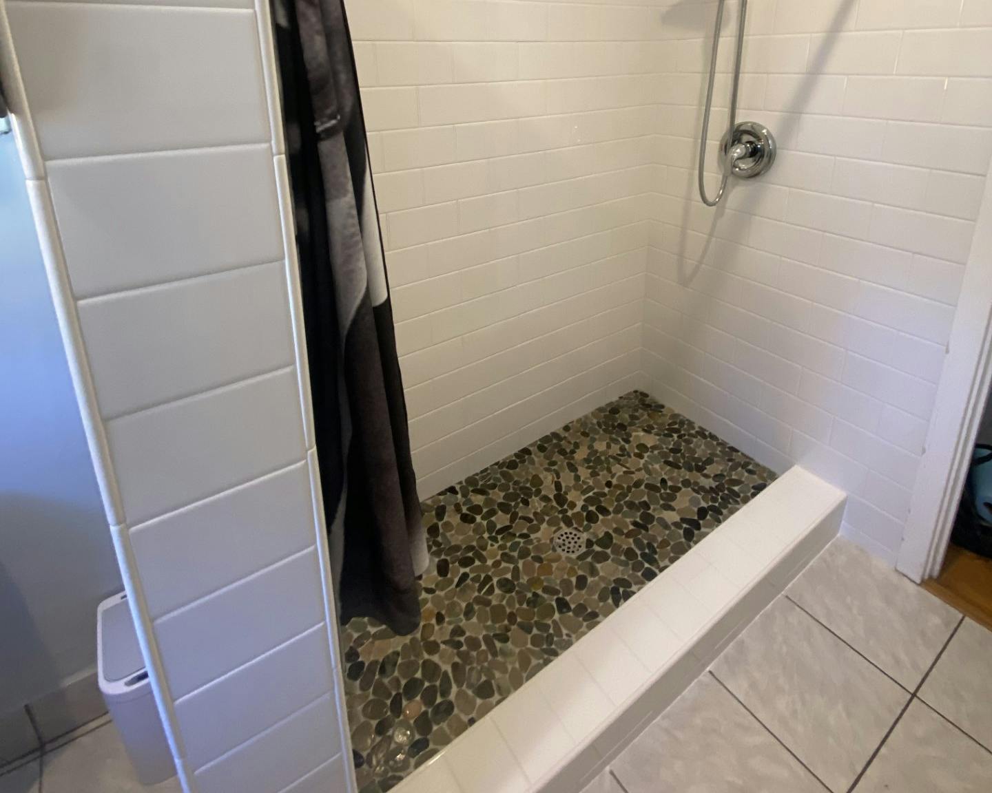 "A modern bathroom corner featuring a walk-in shower with white horizontal tiling on the walls and a pebble stone floor. A stainless steel handheld showerhead is mounted on the right wall. To the left, a white folding laundry hamper stands next to a draped dark shower curtain. The foreground shows a glimpse of a white bathtub edge and gray floor tiles.